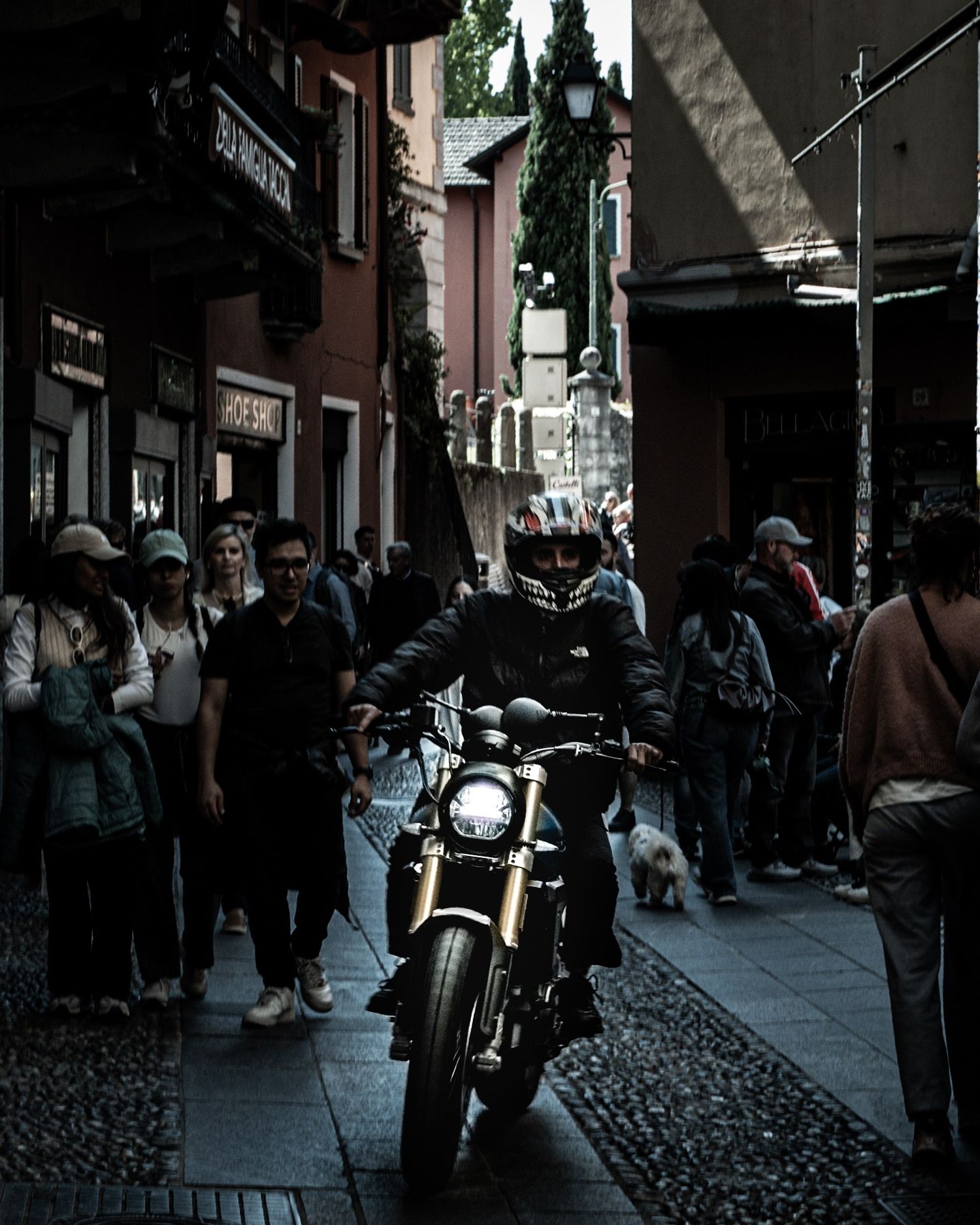 A motorcyclist in a helmet rides through a narrow cobblestone street lined with pedestrians and shops.