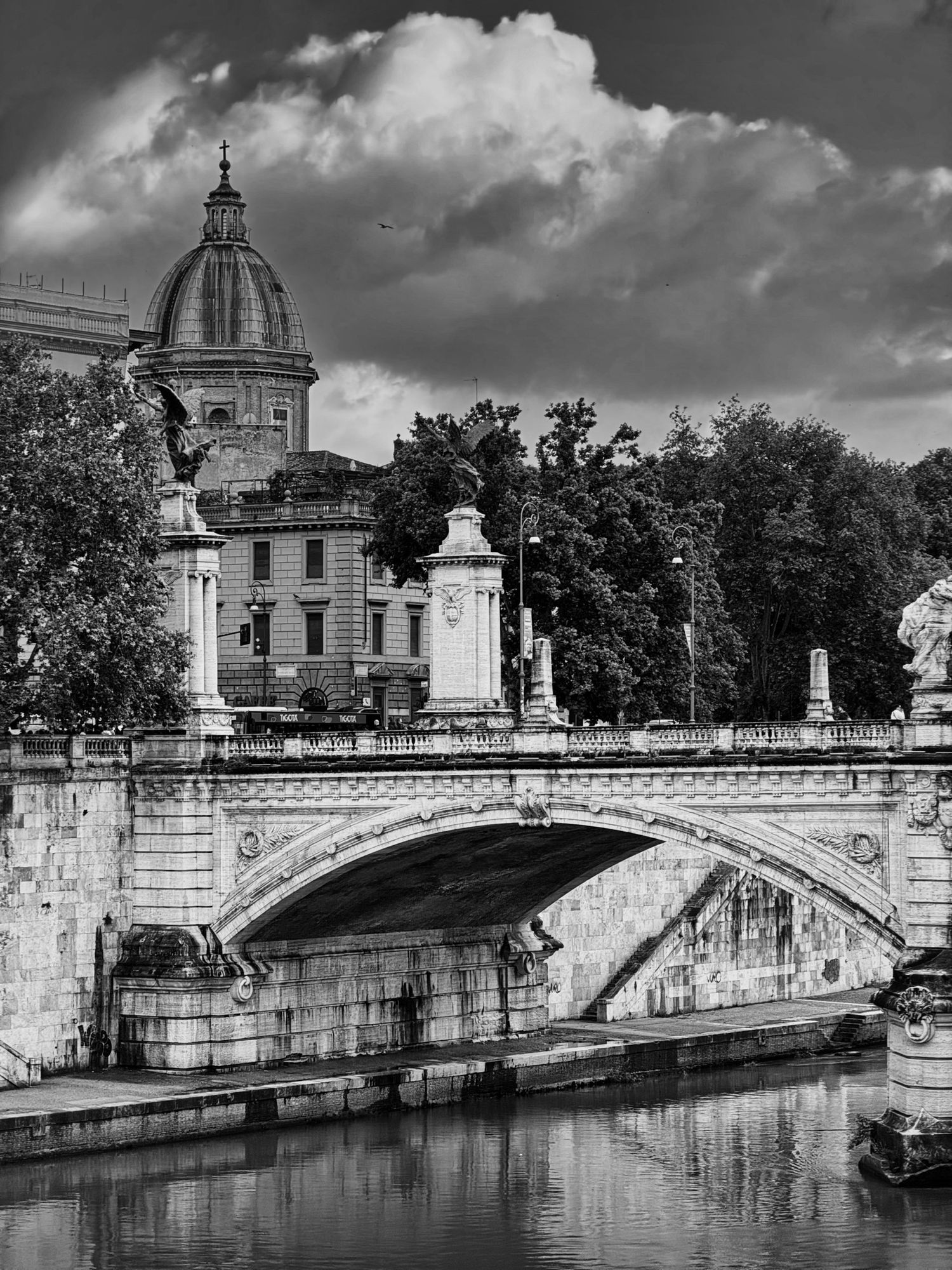 Black and white photo of an ornate stone bridge over a river with a domed building and trees in Rome, Italy.