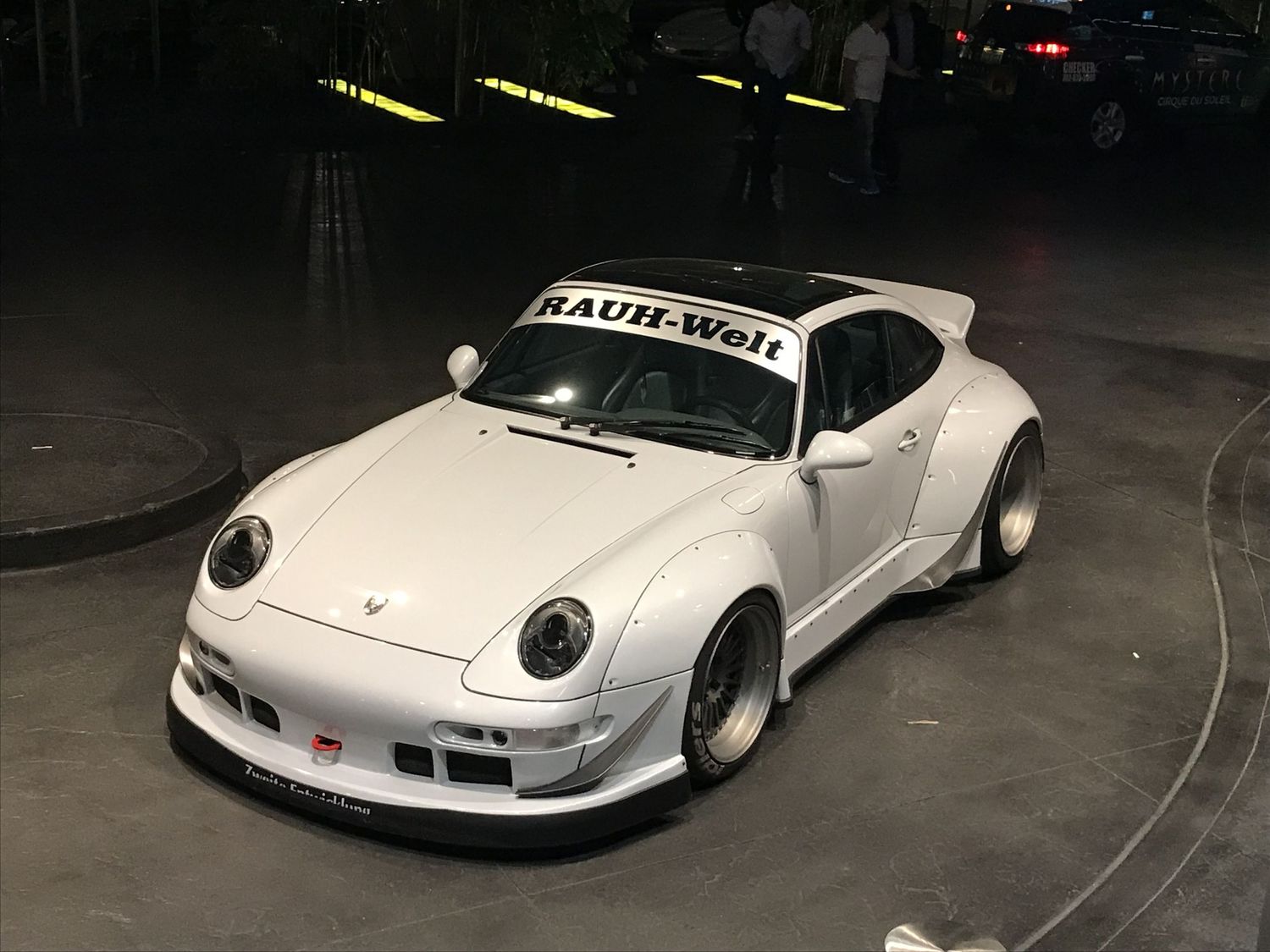 White widebody Rauh-Welt Begriff RWB Porsche 993 with aggressive body kit displayed at an indoor car show.