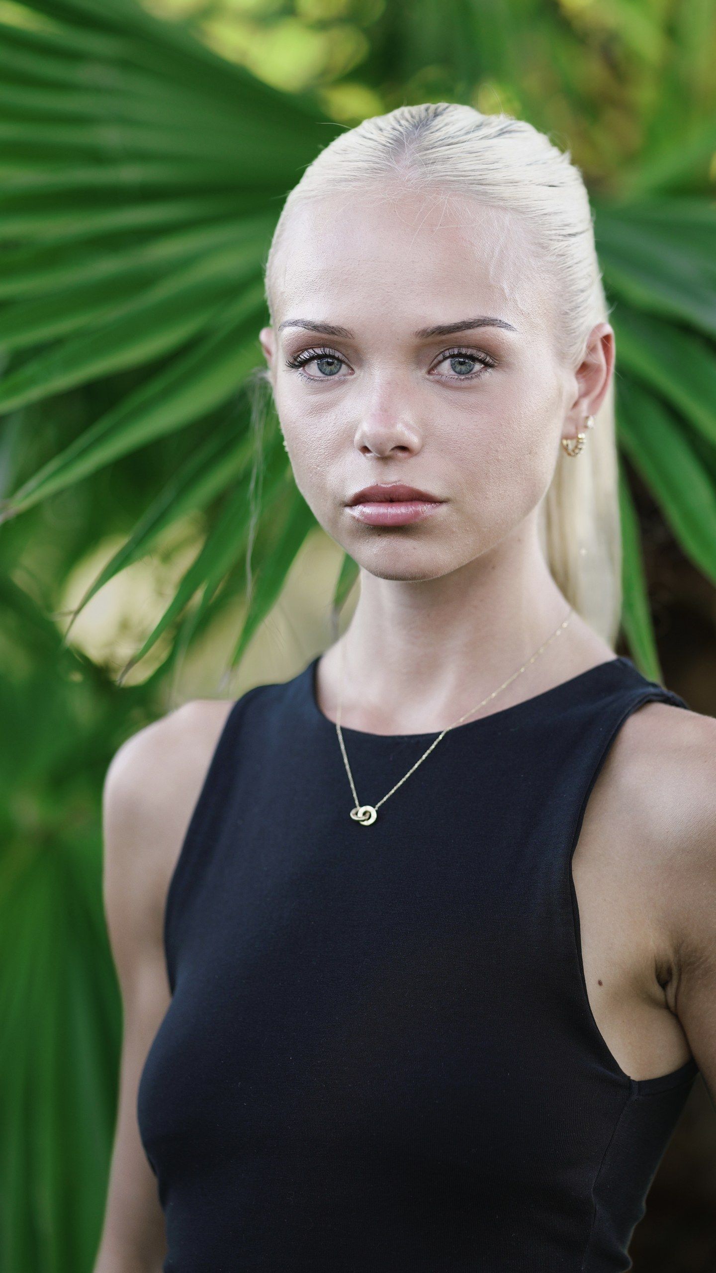 Young blonde woman in black sleeveless top with gold necklace posing outdoors near tropical palm leaves.