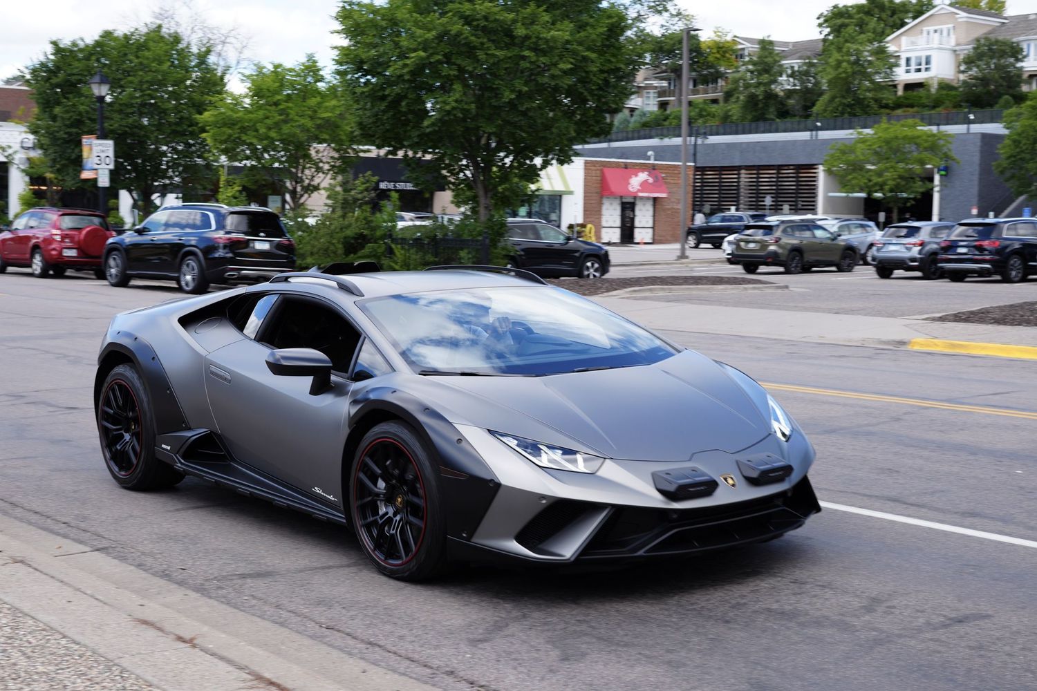Matte gray Lamborghini Huracan Sterrato supercar driving on a suburban street lined with trees.