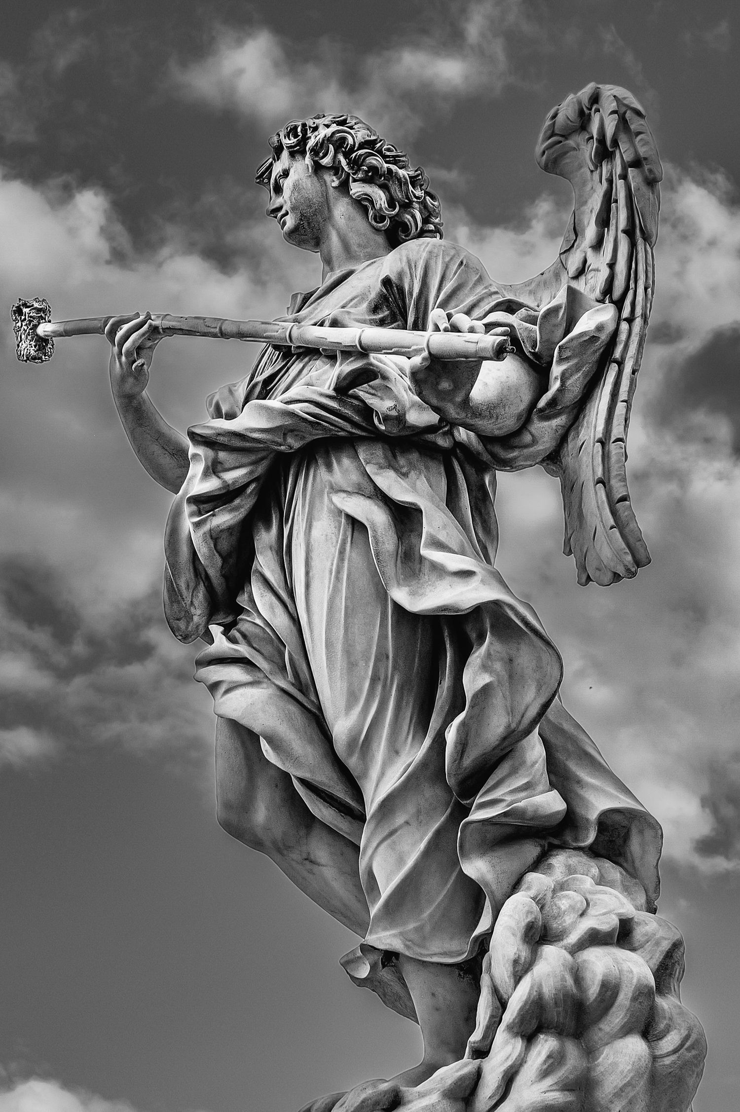 Black and white photo of an angel statue holding a staff, set against a cloudy sky in Rome.