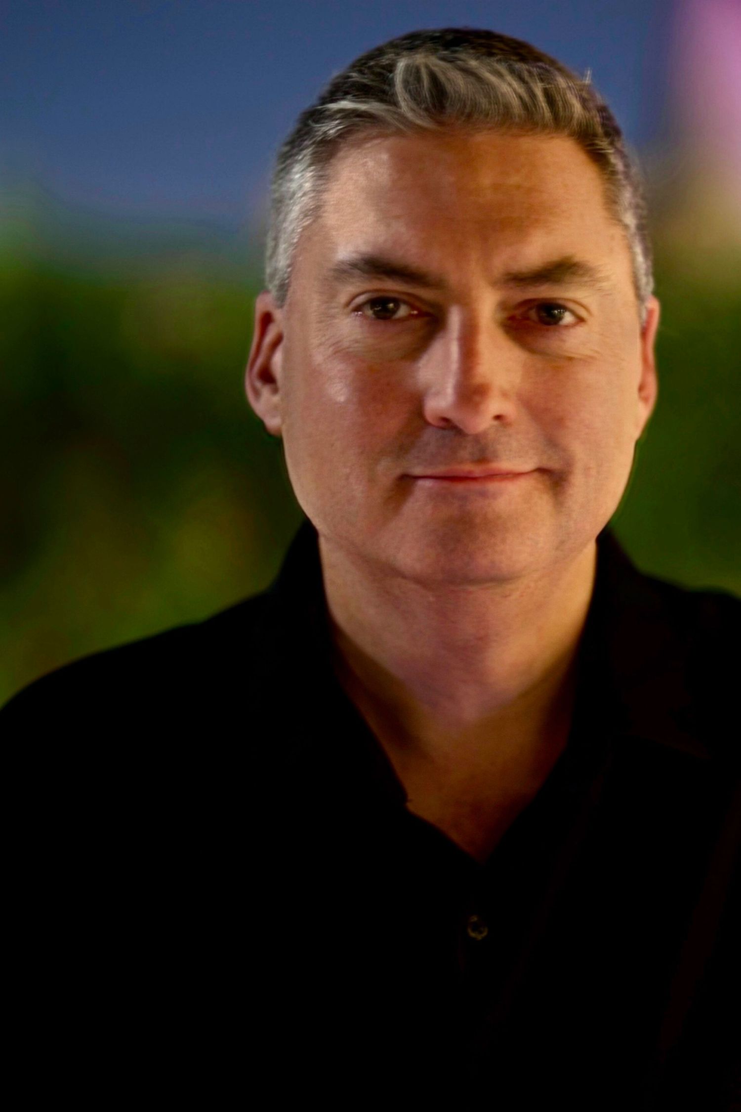 Middle-aged man with gray hair wearing a black polo shirt, posing for a professional portrait outdoors at dusk.