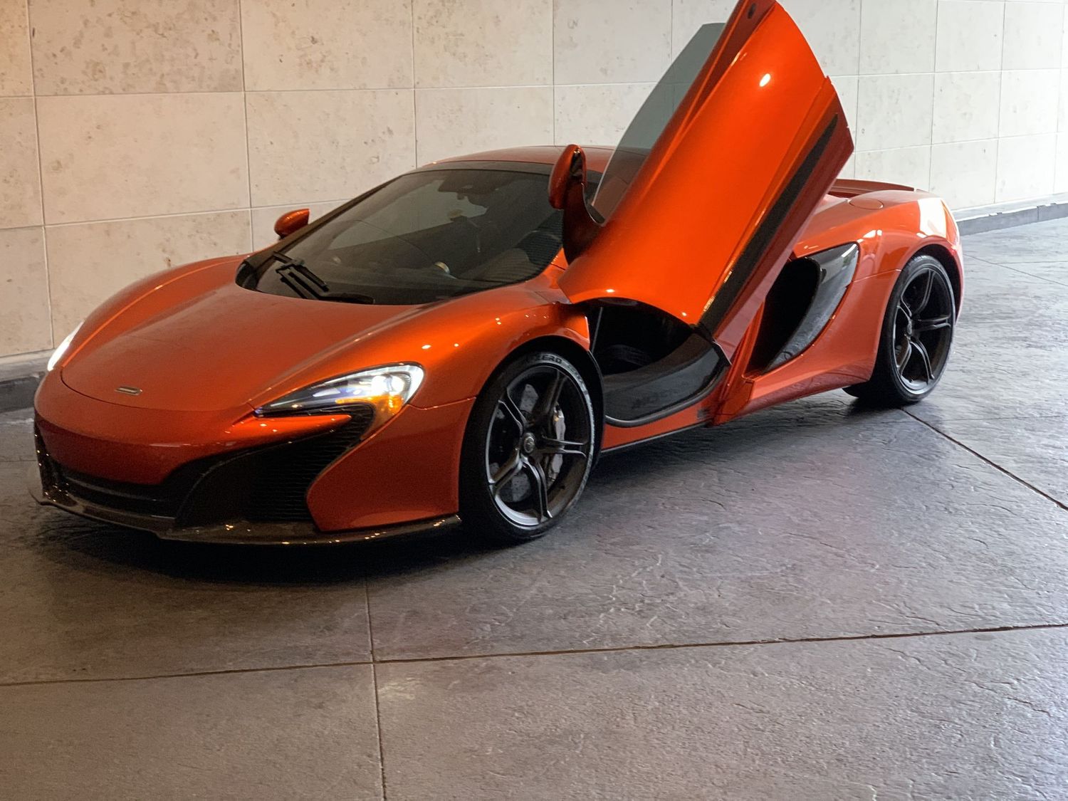 Orange McLaren 650S supercar with dihedral doors open wide, parked on marble floor inside a luxury showroom.