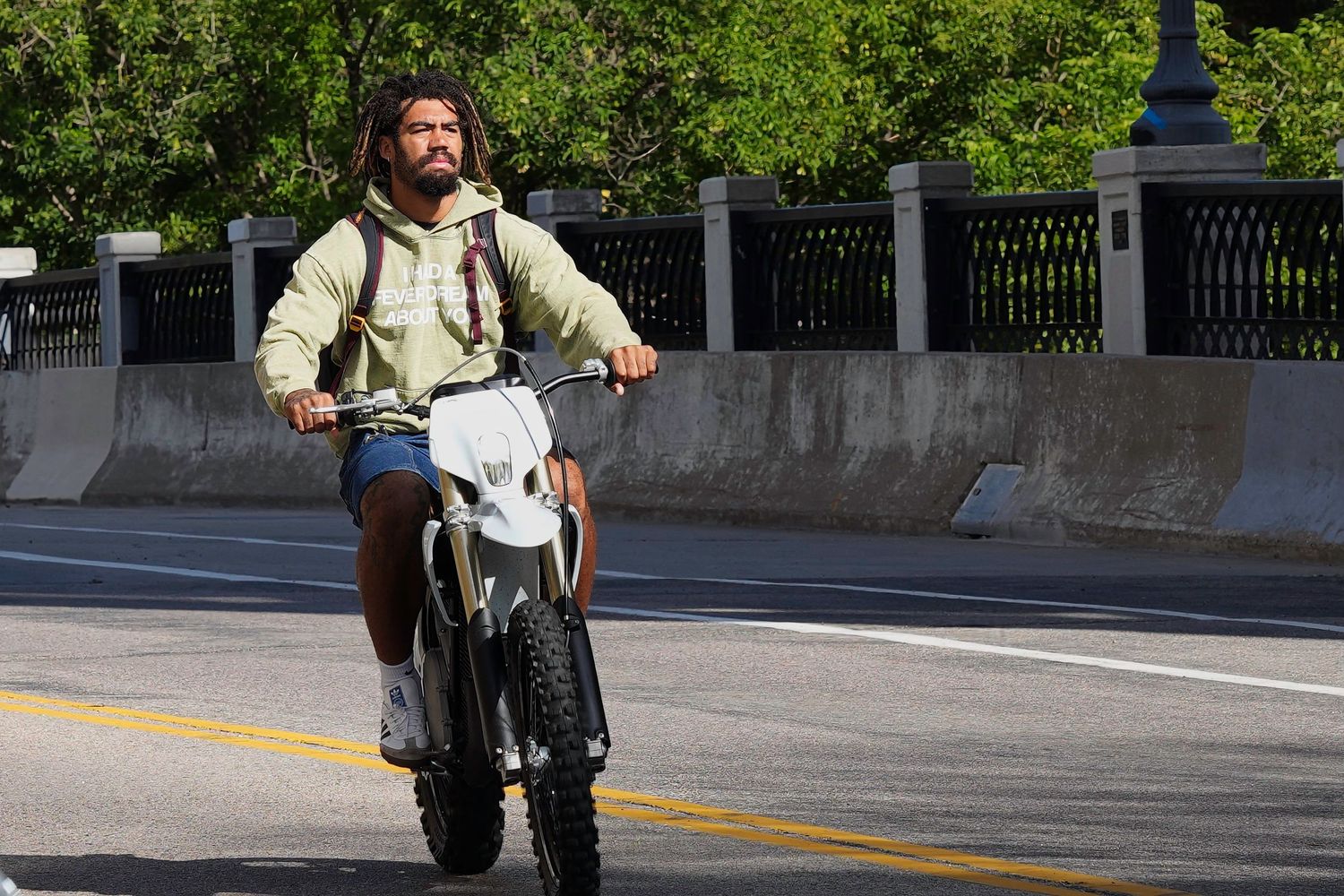 Person with backpack riding a motorcycle along sunny urban street with trees.