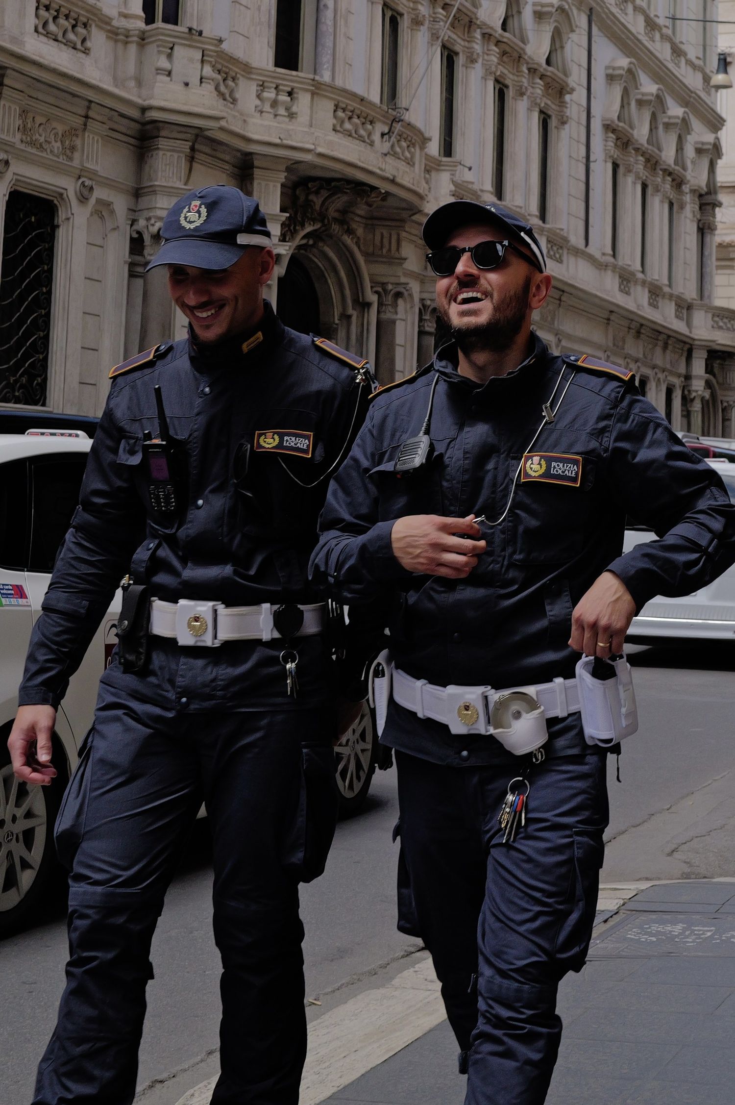 Two smiling Italian police officers in navy uniforms and caps walking together on a city street.