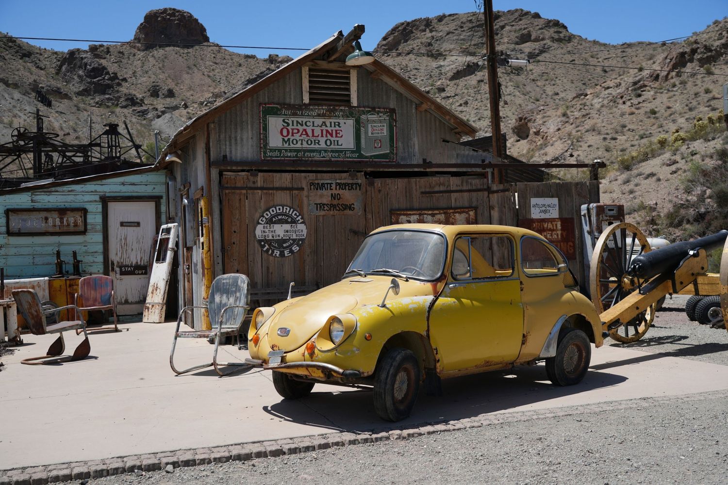 Vintage yellow Subaru 360 parked outside a rustic desert gas station with weathered wooden facade in arid landscape.