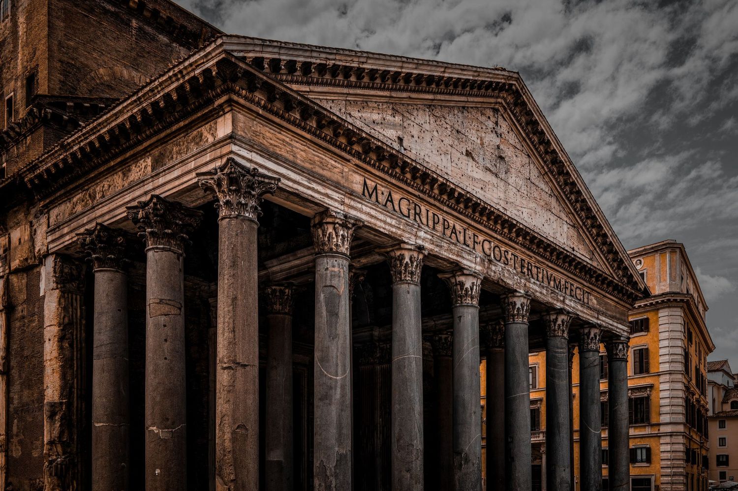 The ancient Roman Pantheon facade with towering Corinthian columns and Latin inscription under dramatic cloudy sky.