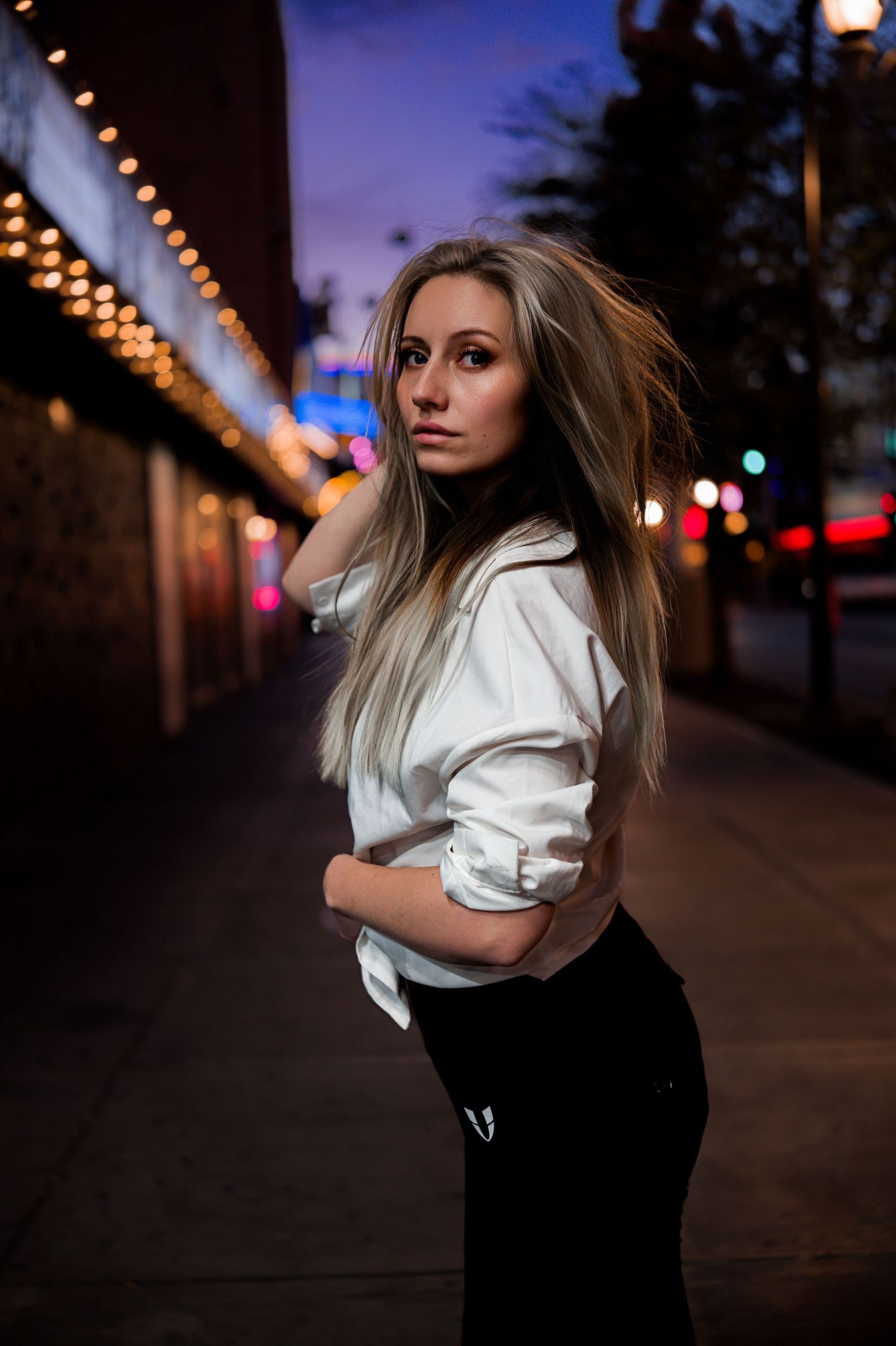 A person in casual attire poses on a city street at dusk with string lights in the background.