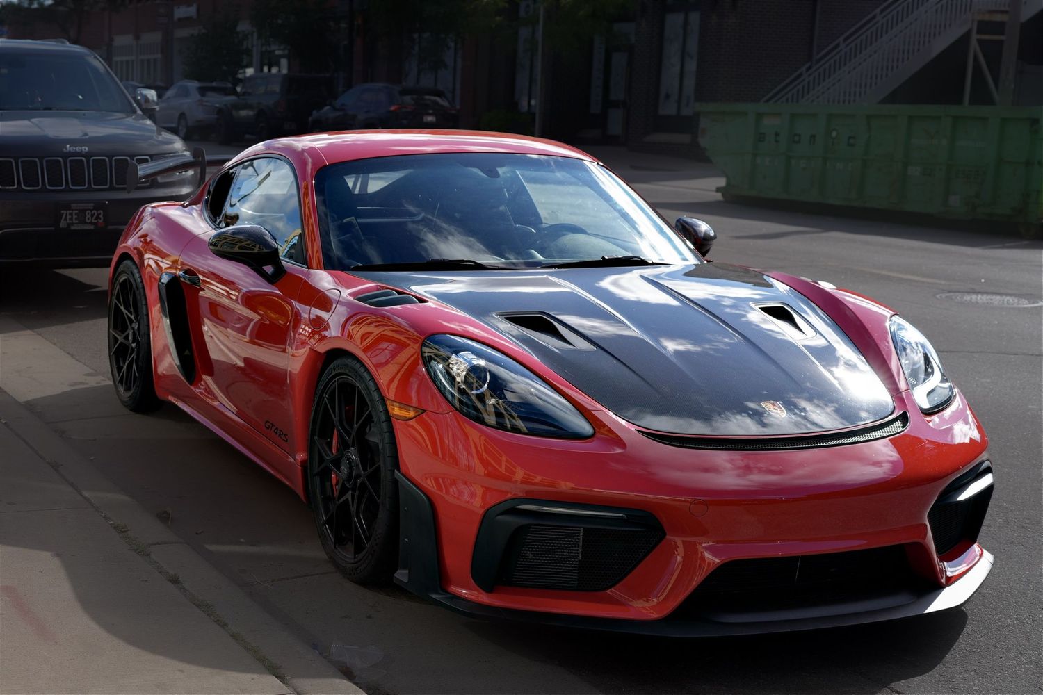 Red Porsche 718 Cayman GT4 RS with carbon fiber hood and black wheels parked on a city street.