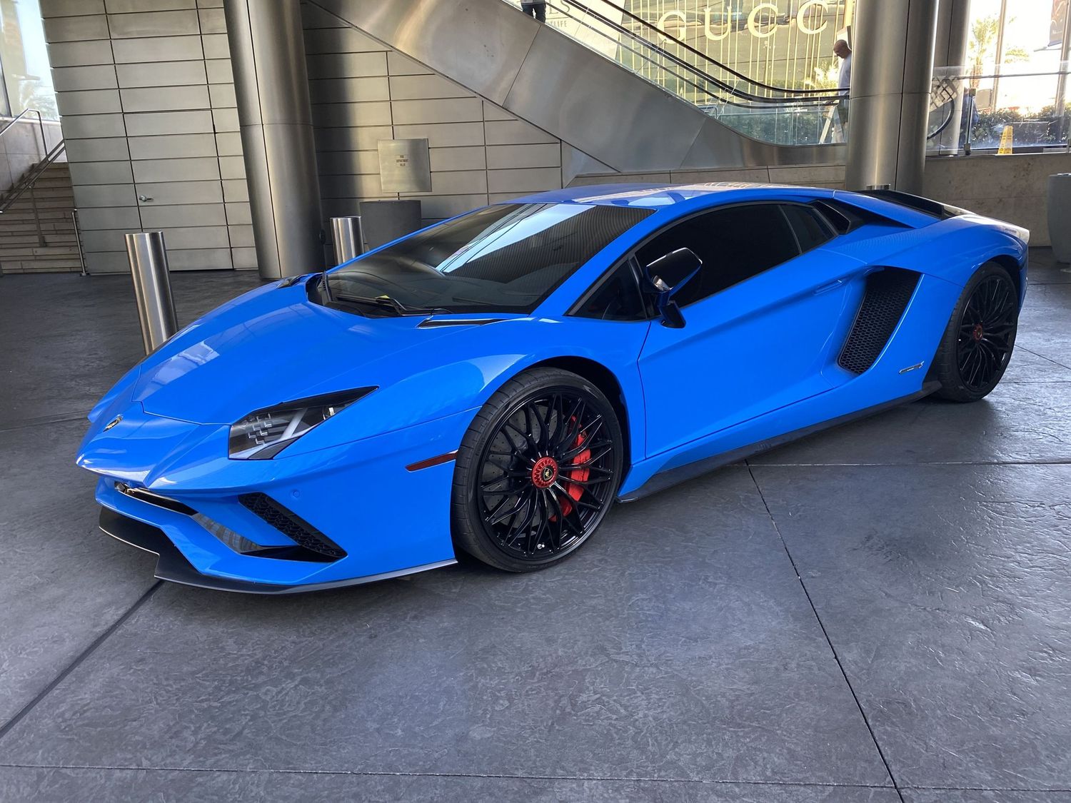 Bright blue Lamborghini Aventador supercar parked outside a modern building with black wheels and red brake calipers.