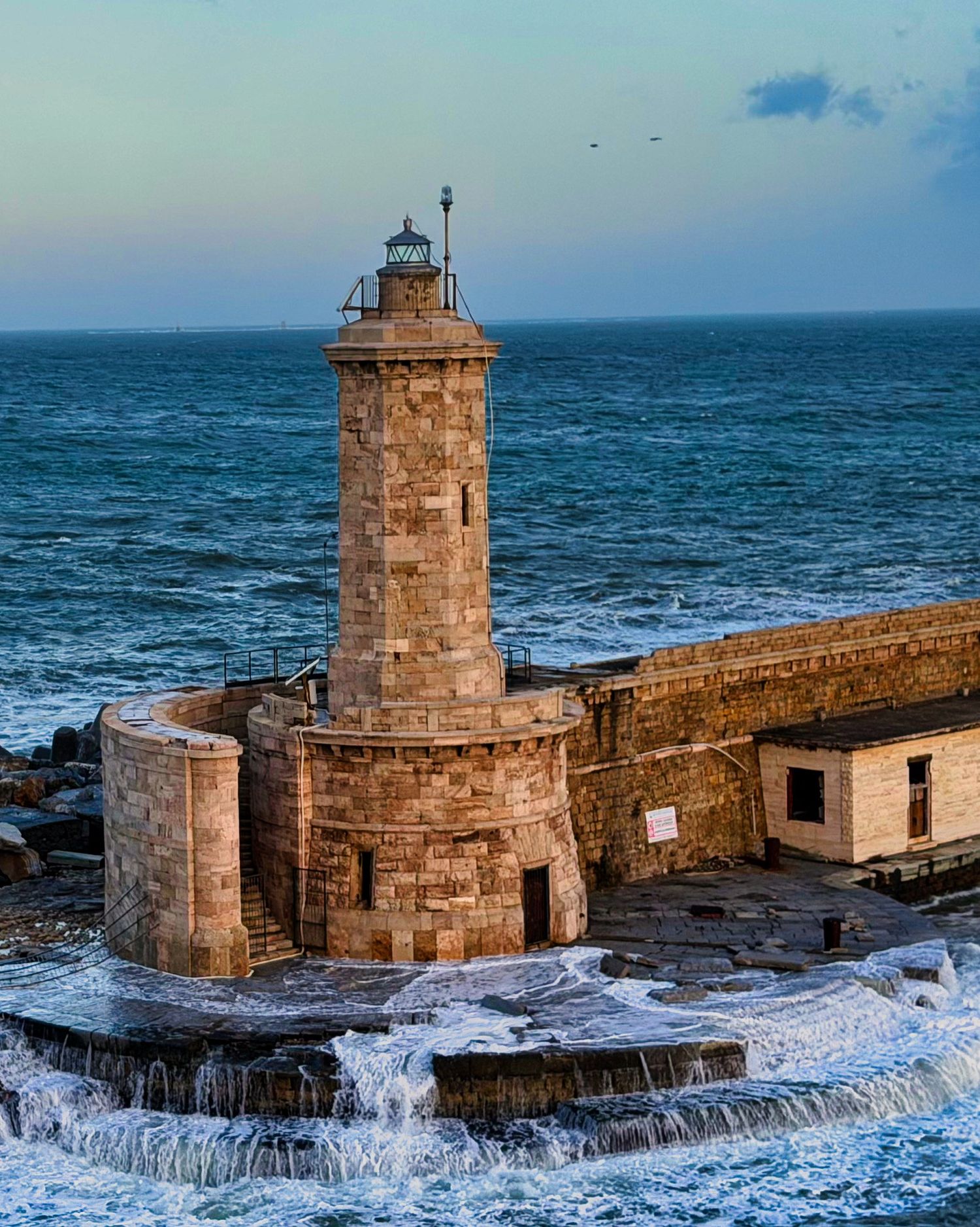 Historic stone lighthouse on rocky coastal pier with crashing waves and blue ocean horizon at dusk.