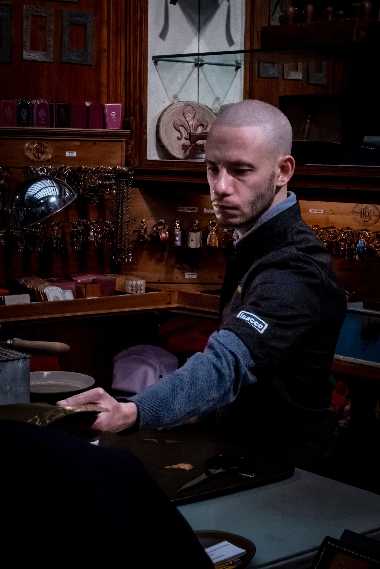 Bald young man sitting at a wooden antique shop counter, surrounded by jewelry and collectibles on display shelves.