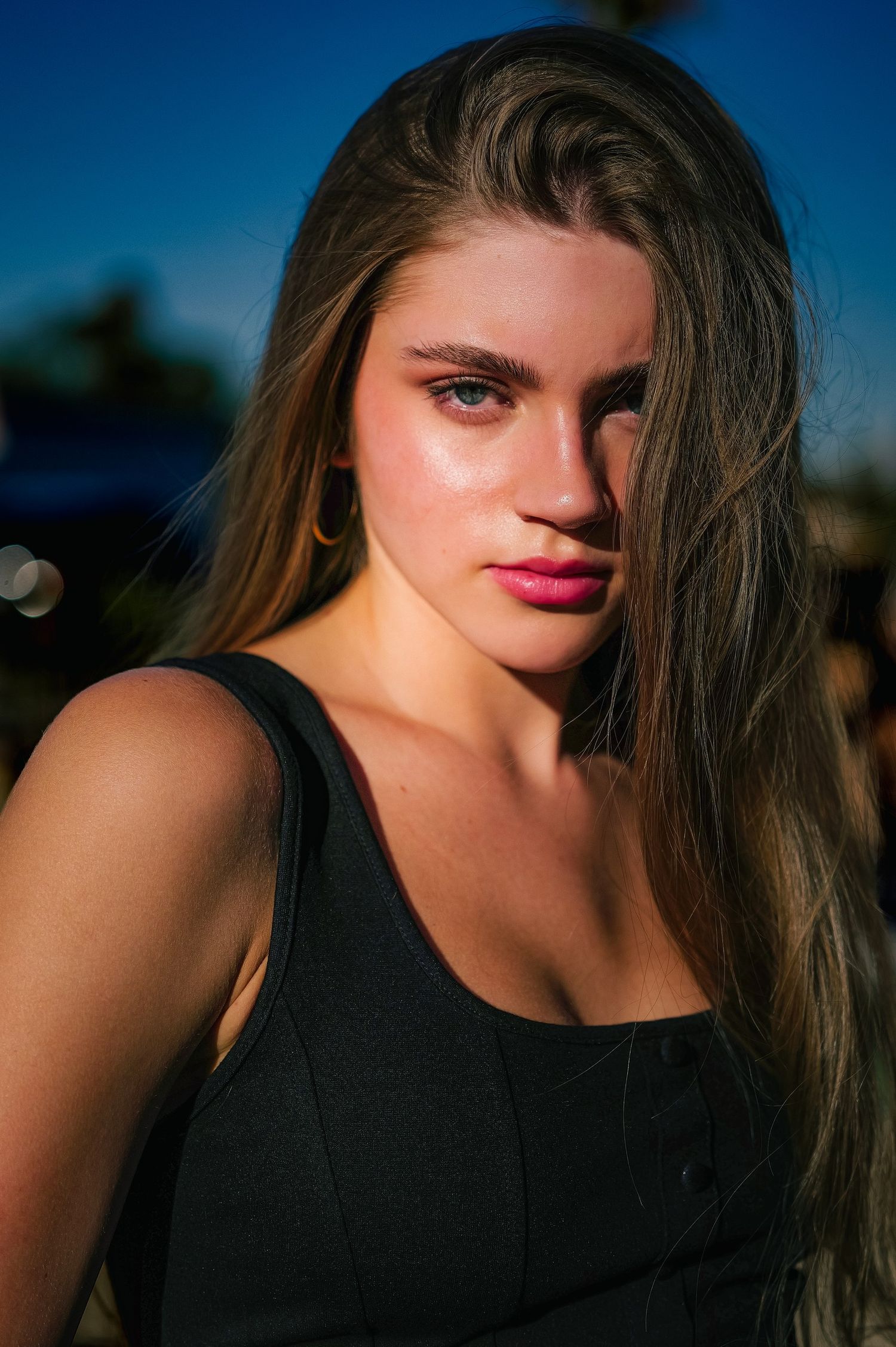Young woman with long brown hair wearing a black tank top poses outdoors in warm golden sunlight.