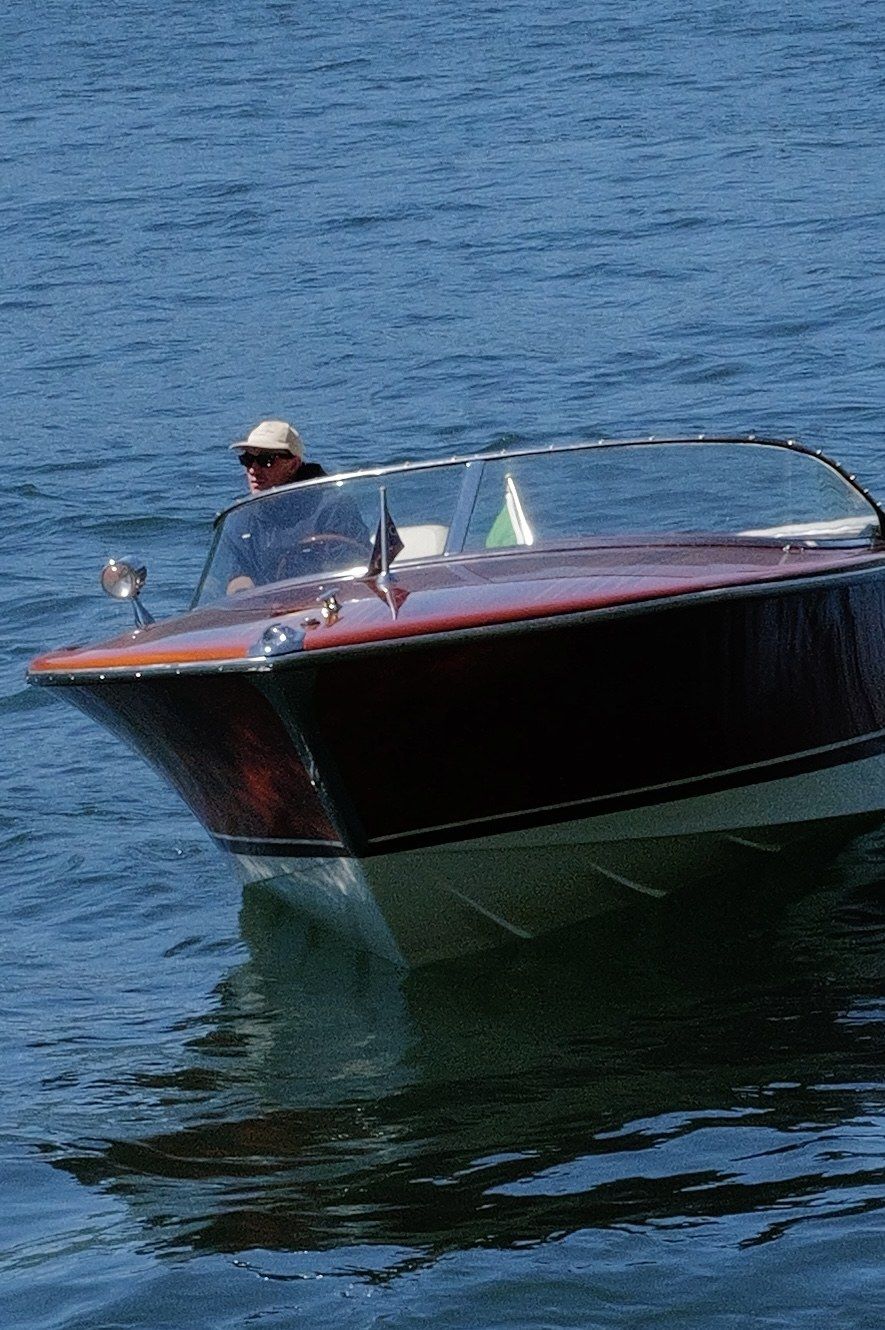 Classic wooden Riva speedboat gliding across calm blue waters with a driver wearing a hat.