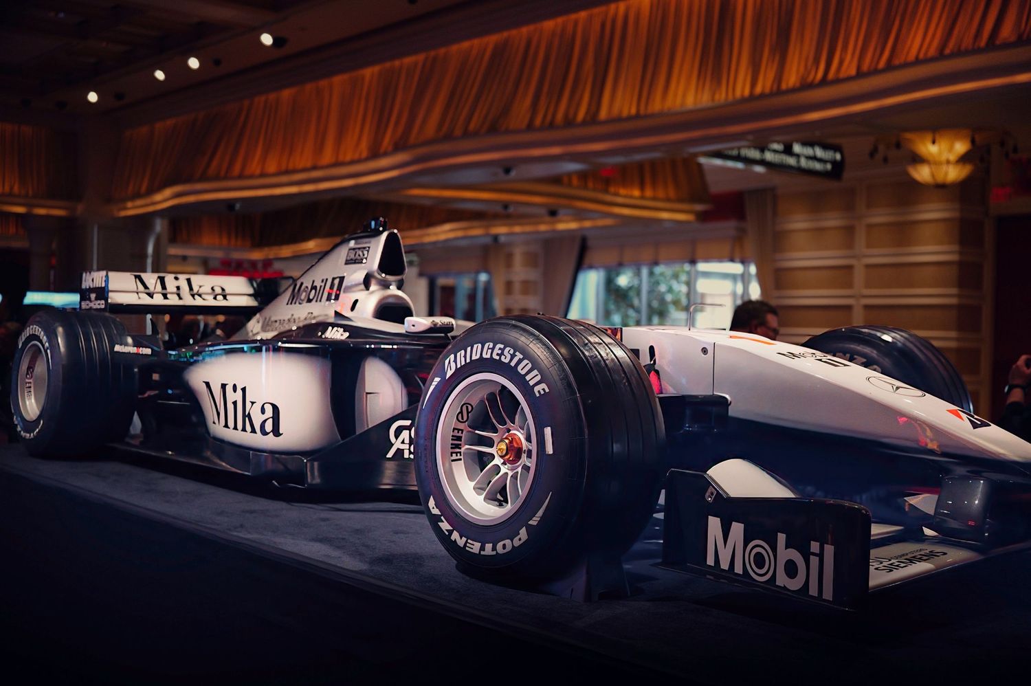 McLaren Formula 1 race car with Mika and Mobil sponsorship displayed indoors under dramatic lighting.