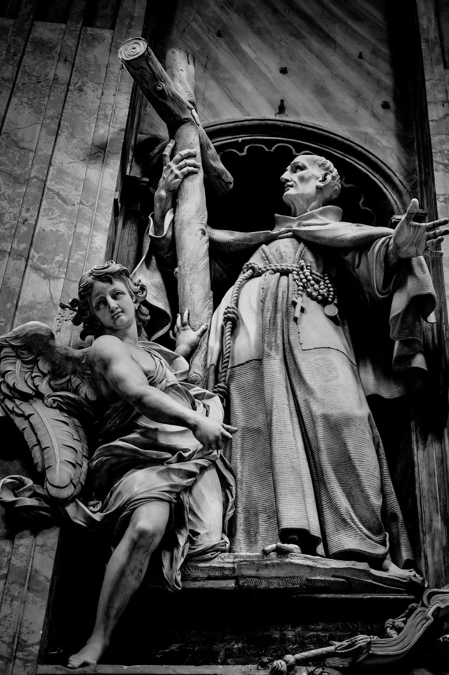 Black and white photo of baroque church statues depicting a saint with an angel holding a cross.