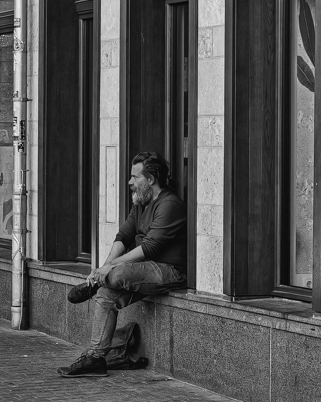 Black and white photo of a bearded man sitting on a ledge outside a building, gazing thoughtfully to the side.