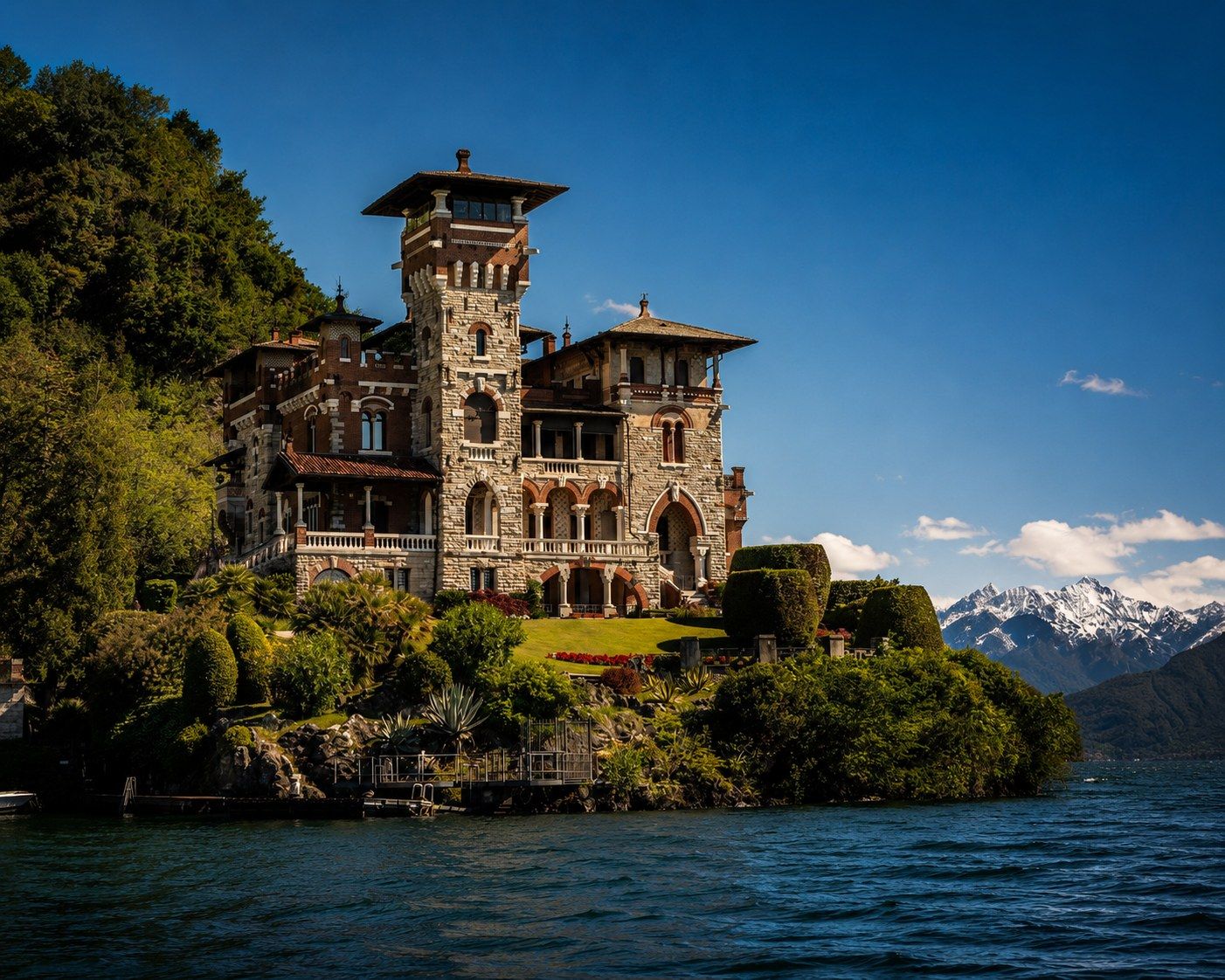 Historic stone castle villa on Lake Como with lush gardens, snow-capped Alps in background under vivid blue sky.