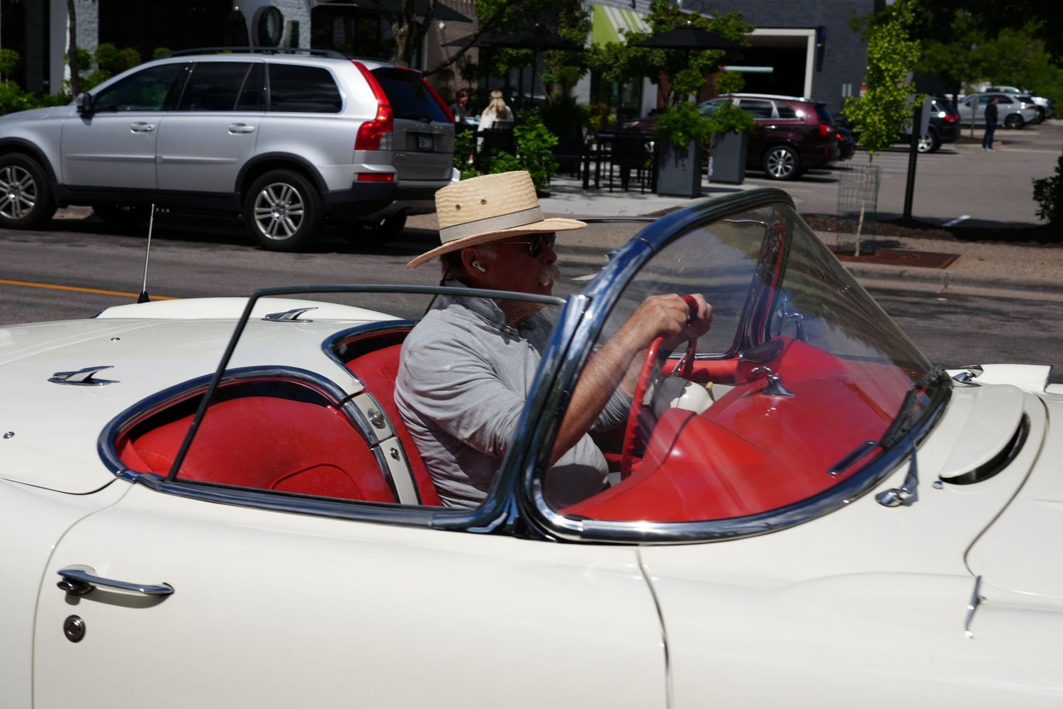 Man in straw hat driving a classic white convertible sports car with red interior on a sunny day.