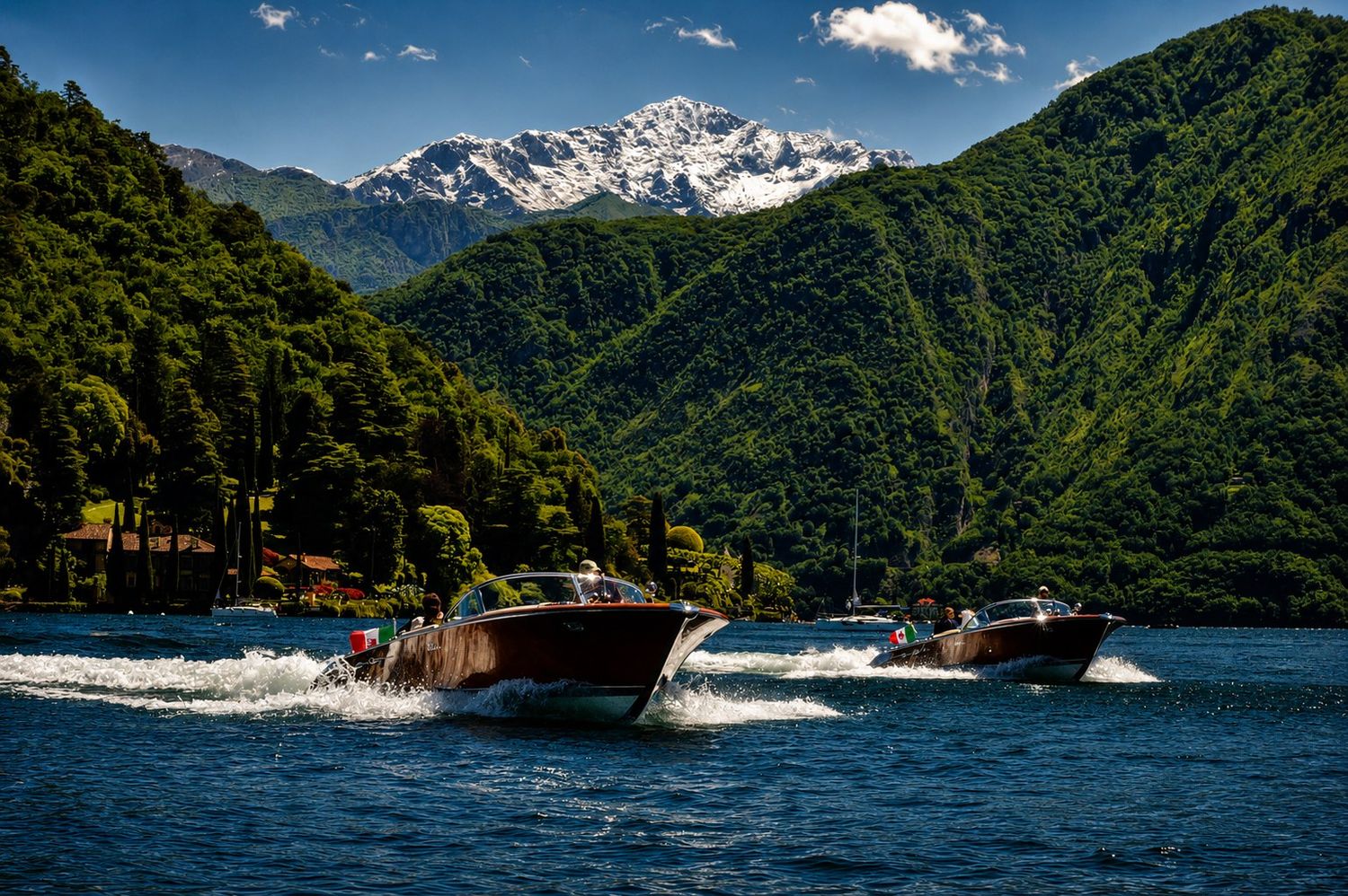 Two classic wooden speedboats race across a scenic alpine lake surrounded by lush green mountains with snow-capped peaks.