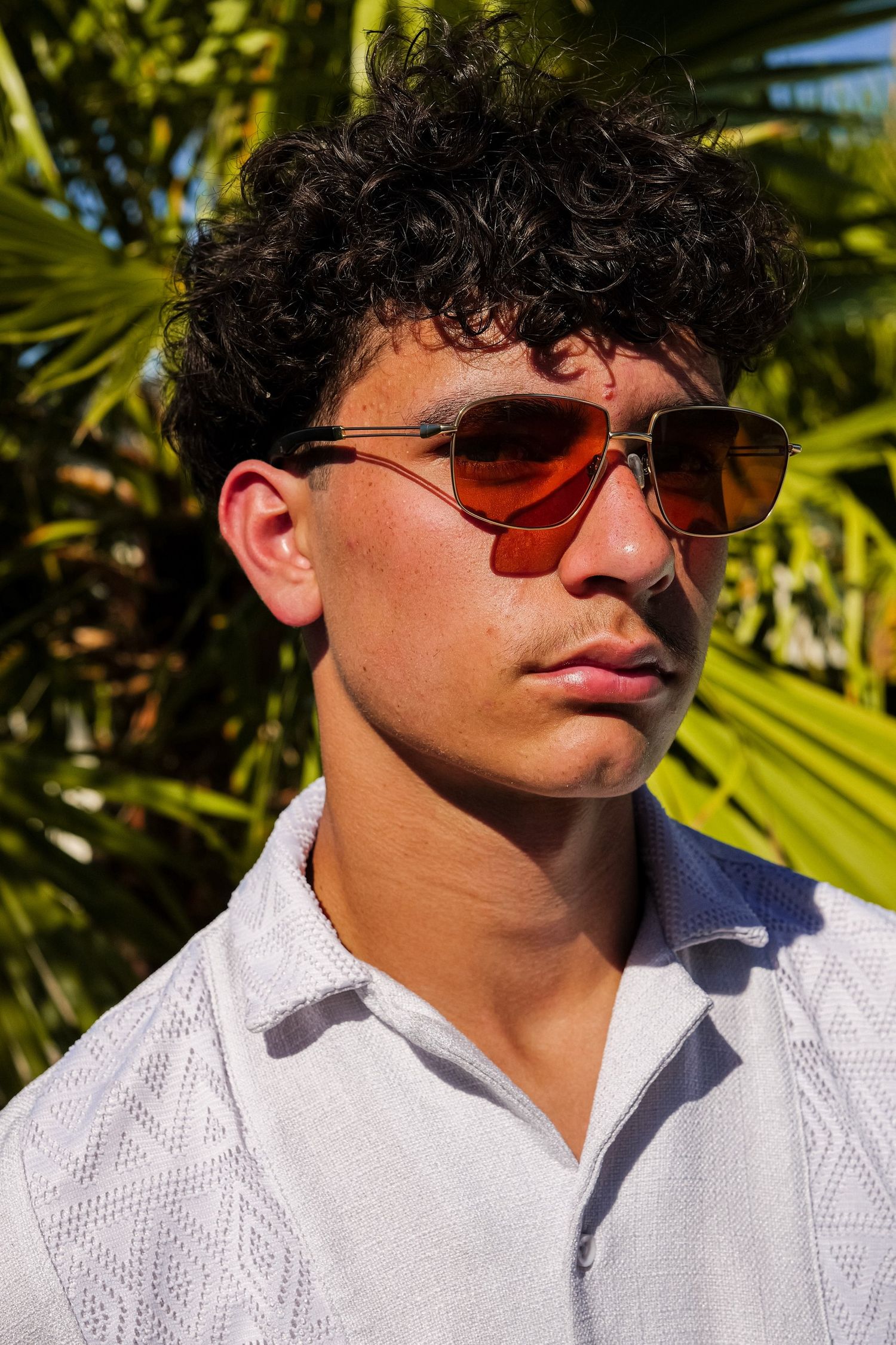 Young man with curly hair wearing red-tinted aviator sunglasses and white shirt outdoors near tropical palms.