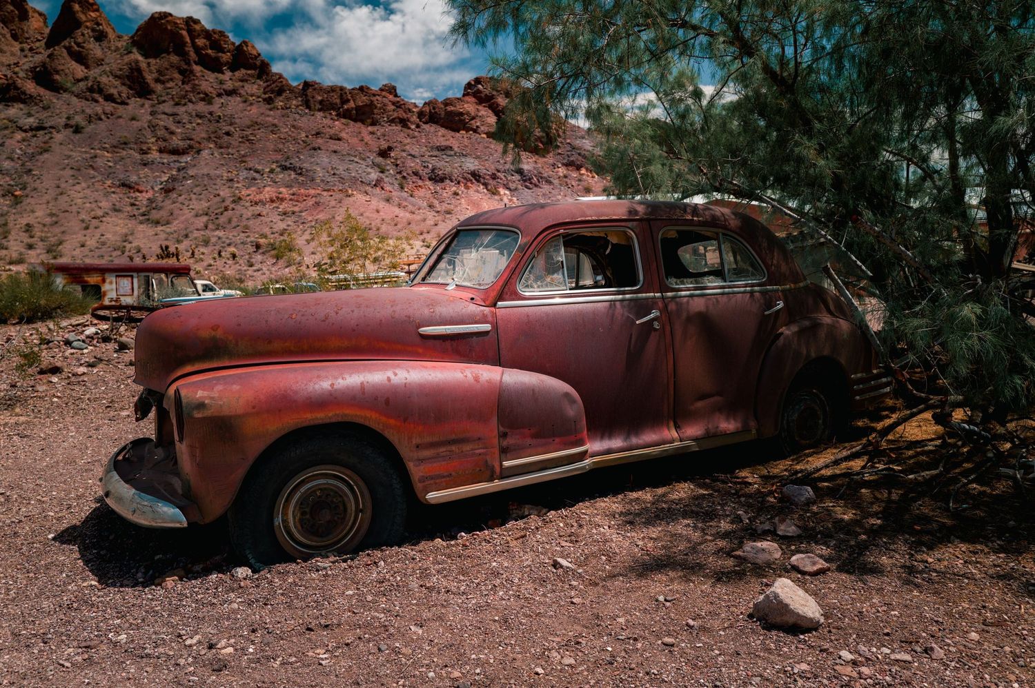 Rusty abandoned vintage car decaying in a desert landscape with red rock formations and sparse trees under a blue sky.