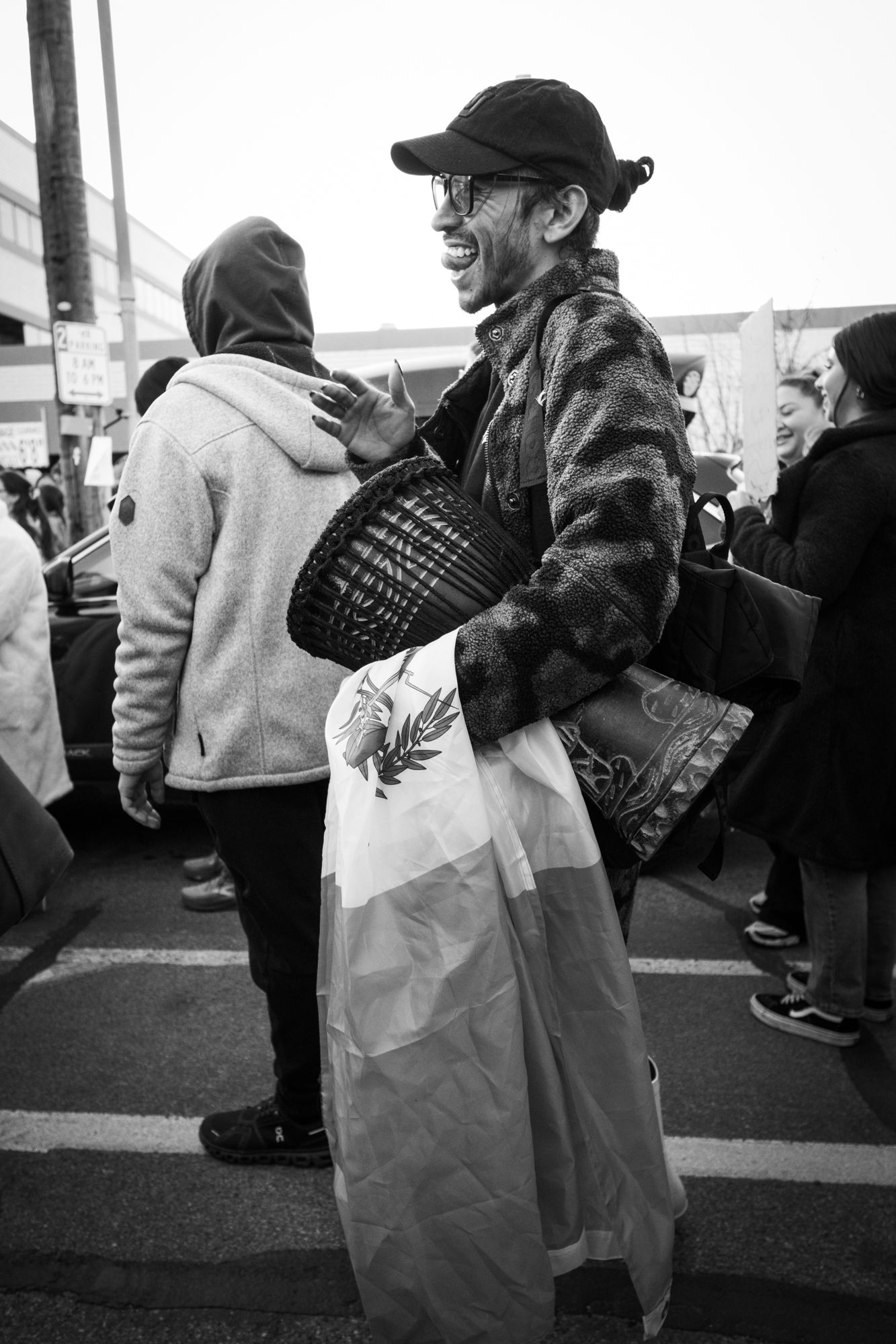 Candid B&W street photography of a joyful drummer with a flag at a public march. Authentic documentary imagery for news and editorial use.