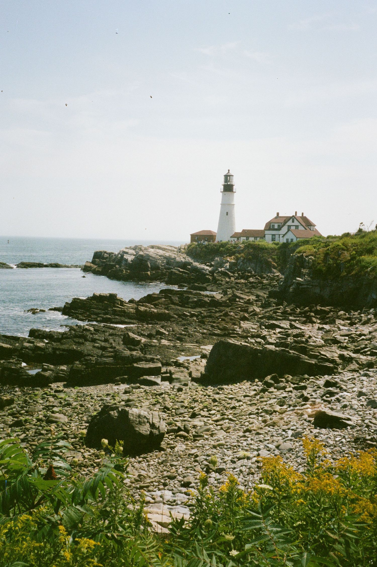 The historic Portland Head Light stands tall over the rocky Atlantic coastline of Cape Elizabeth, Maine, under a soft, hazy summer sky.