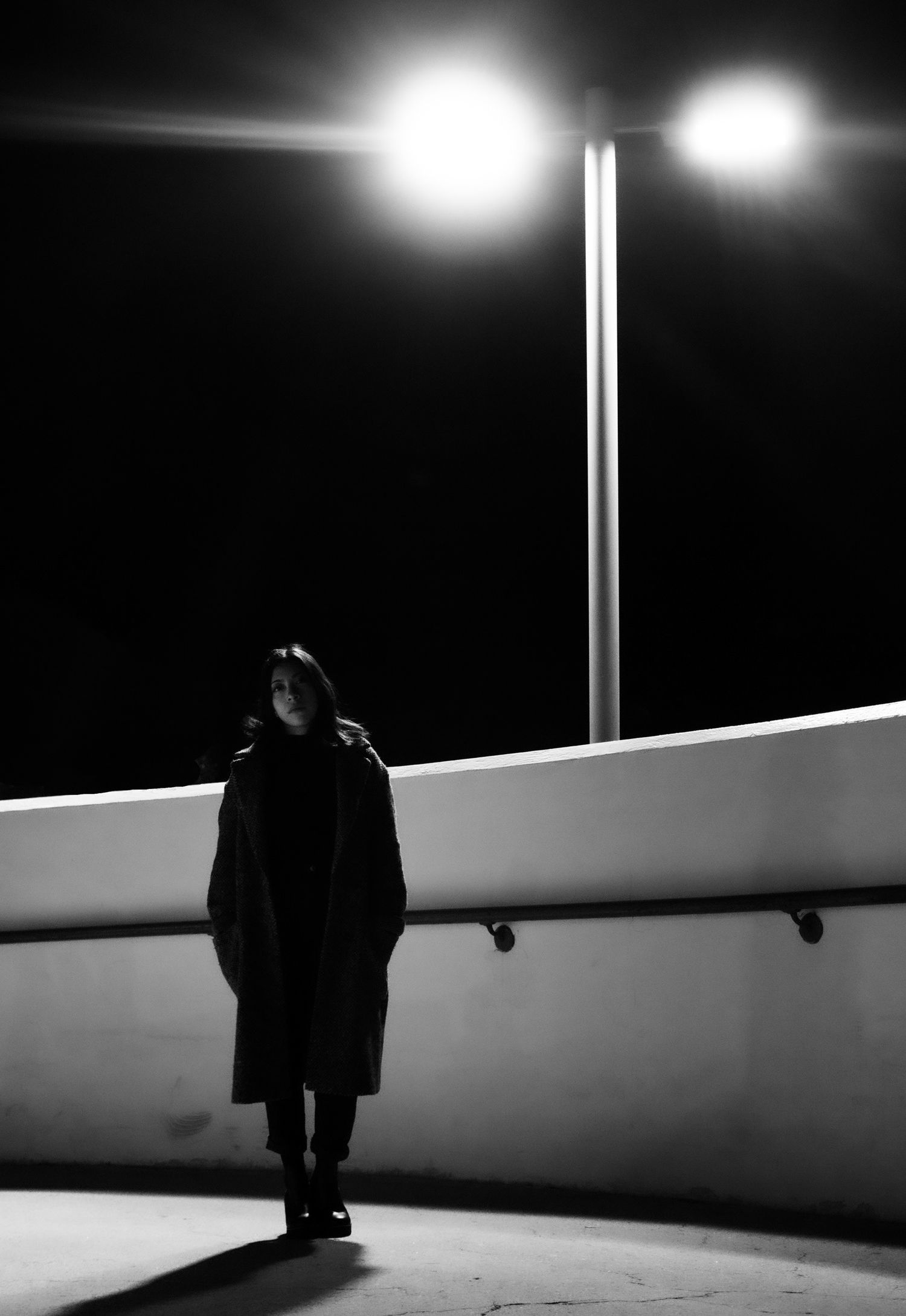 A haunting black-and-white night portrait of a woman standing beneath the glowing lens flare of a street lamp, creating a cinematic noir