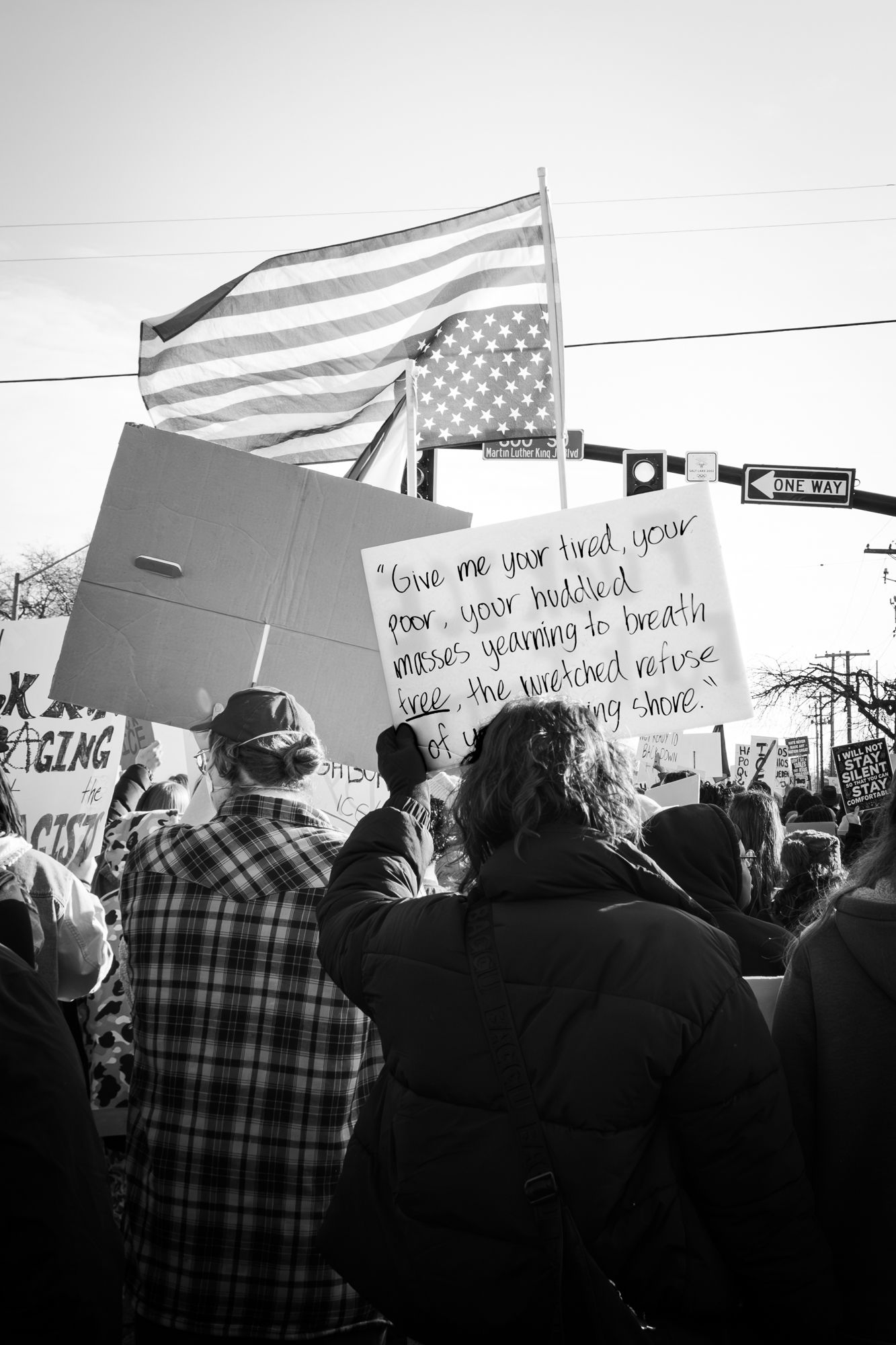 Moving black & white image of a protest. Features an American flag and a sign with the Emma Lazarus poem. Powerful for advocacy or news.