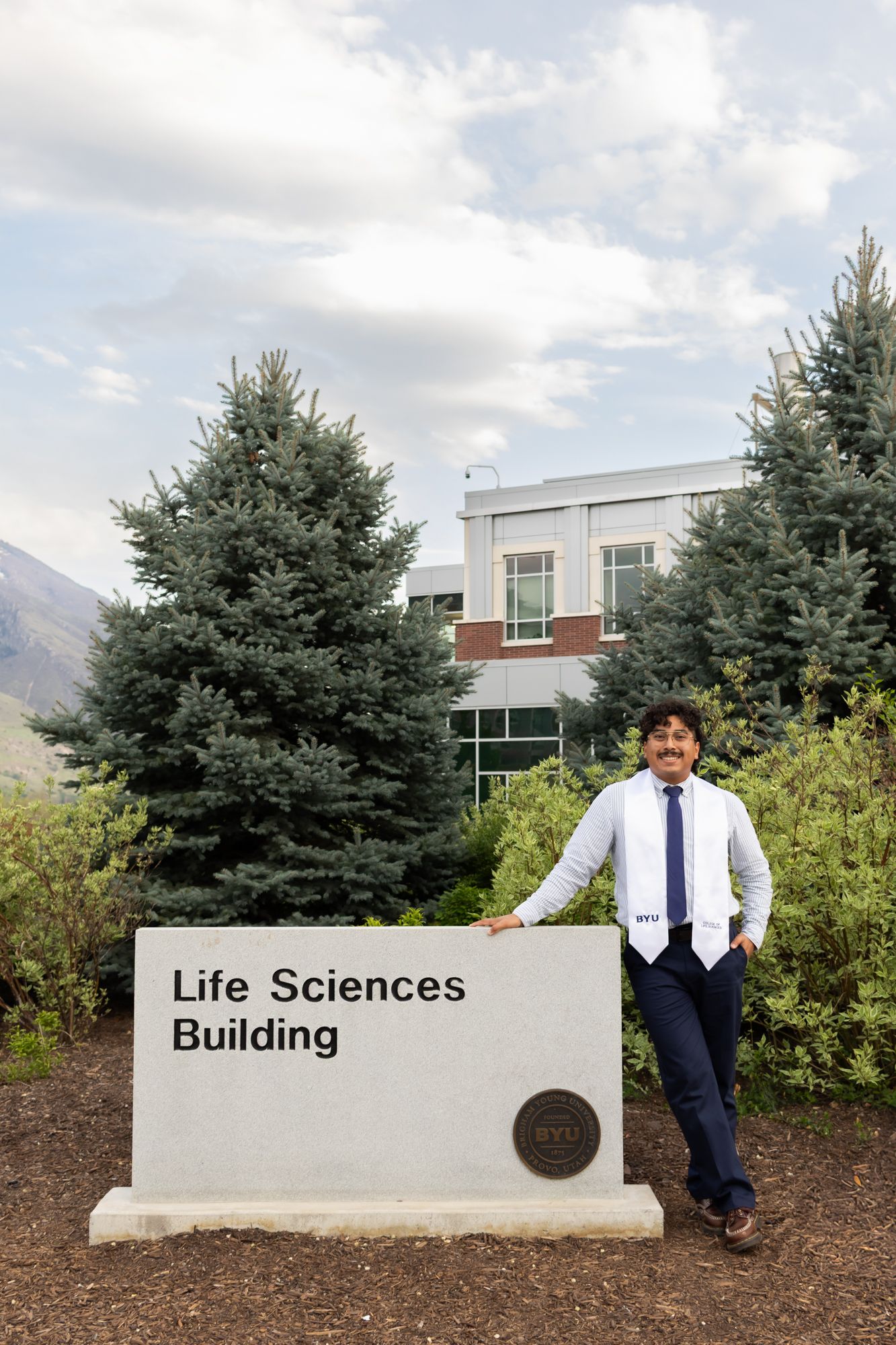 A vertical graduation portrait of a student posing by the BYU Life Sciences Building, capturing a milestone achievement on campus.
