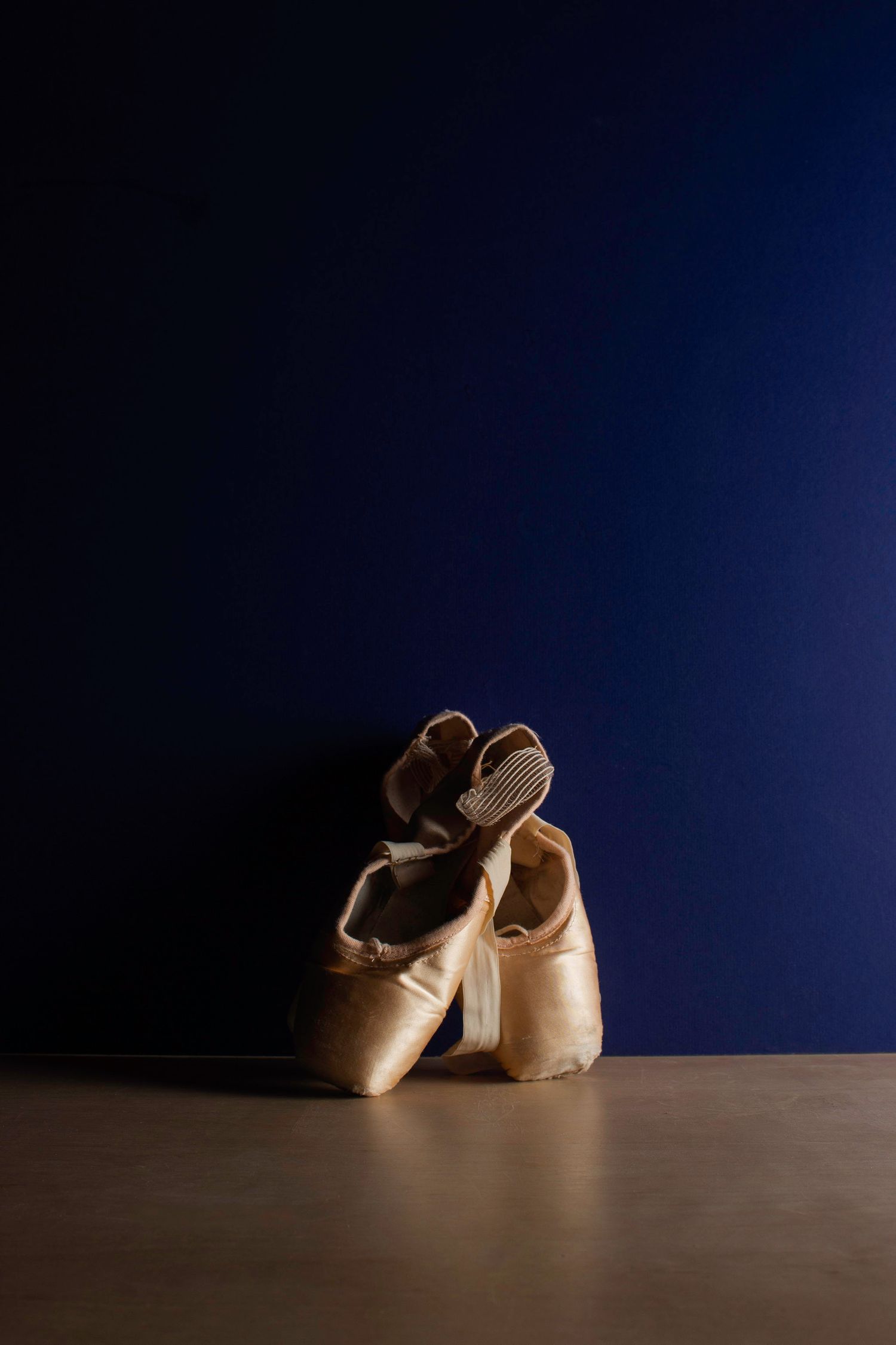 Moody still life of satin ballet shoes against a deep blue background, utilizing dramatic side-lighting to highlight form and texture.