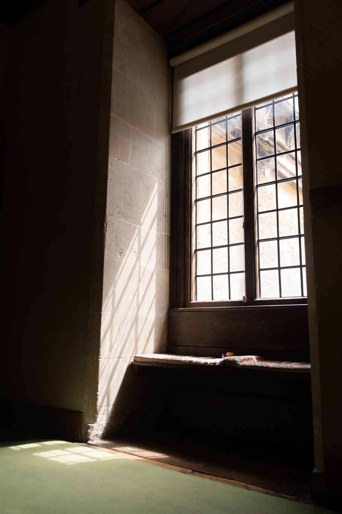 A study of sunlight through a leaded window. Focus on the rhythmic shadow grid cast onto the stone and the quiet, scholarly atmosphere.