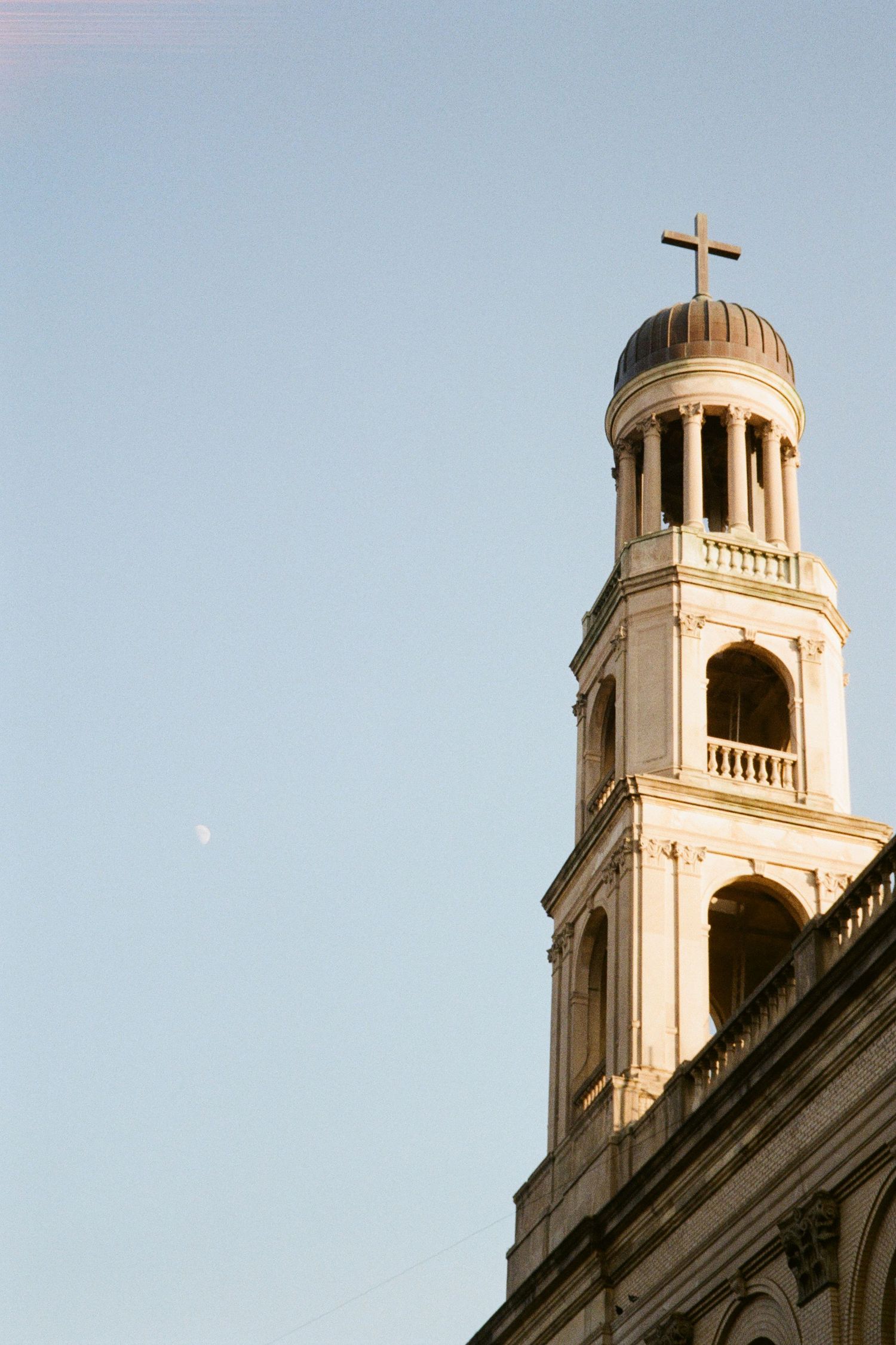 A majestic church bell tower with a cross finial reaches toward a daytime moon, showcasing the historic religious architecture of NYC.