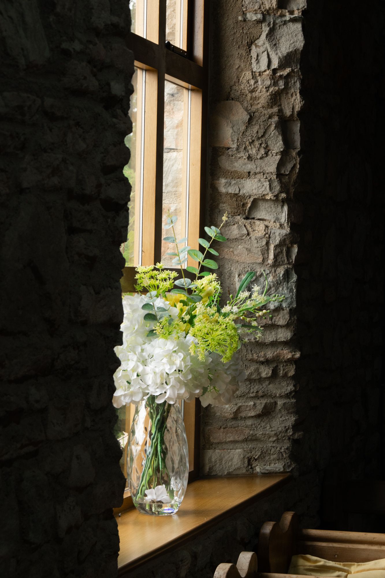 A study of a floral arrangement in a deep stone window sill. Focus on the soft textures against the rugged masonry.