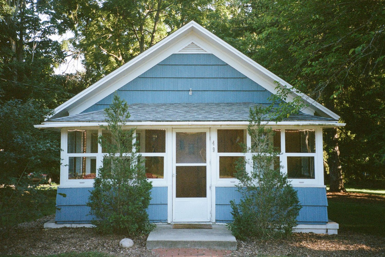 A cozy blue lakeside cottage with a sunroom stands nestled among trees, capturing the peaceful summer retreat vibe of Lake Michigan.