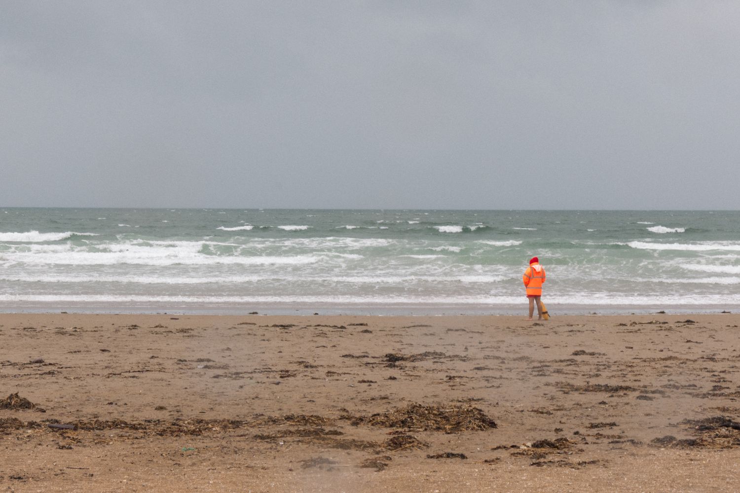 Atmospheric beach study. Focus on the singular orange figure against the rhythmic, horizontal wash of the gray Atlantic. Solitary scale.