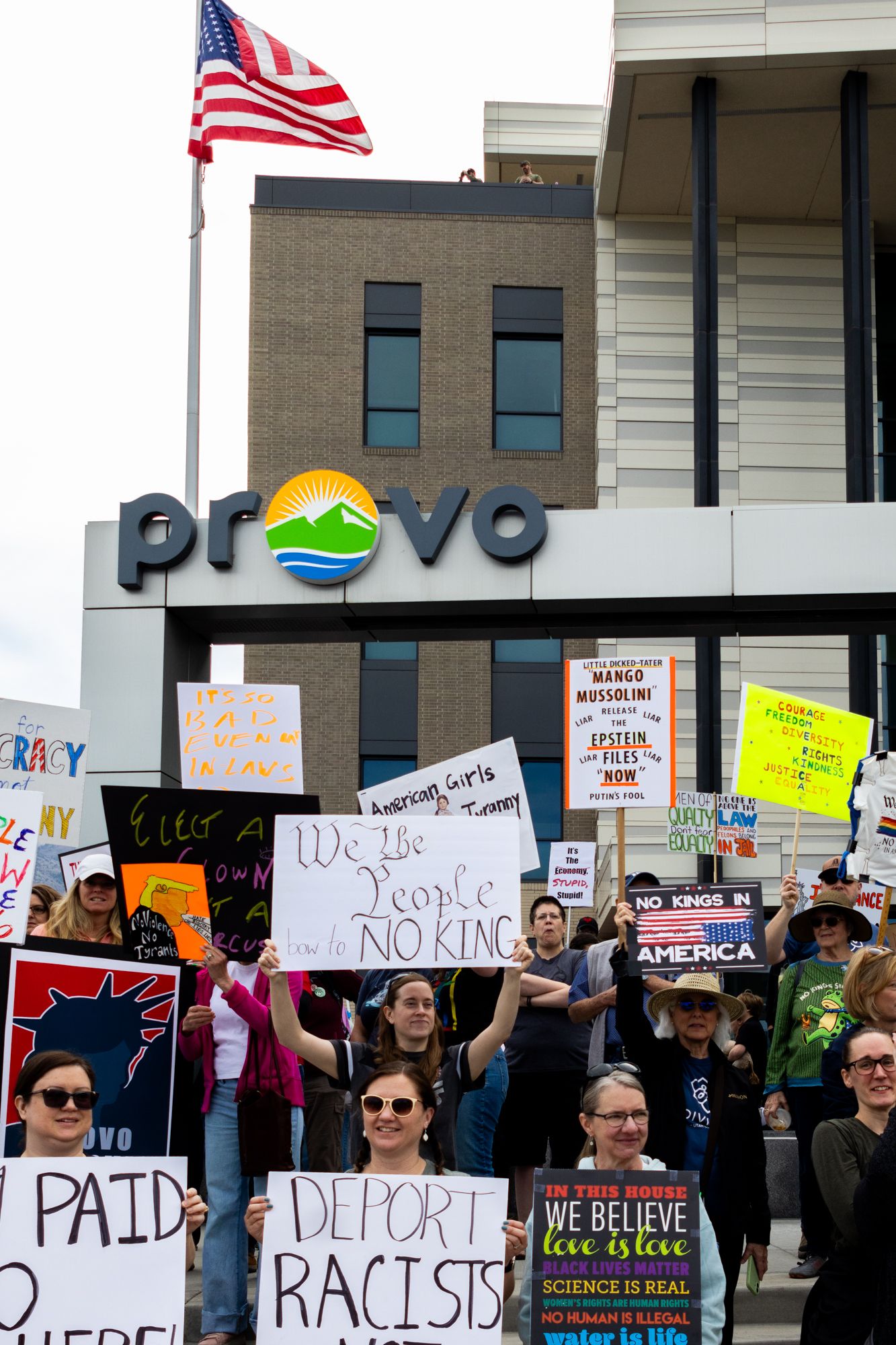A vertical documentary shot of protestors holding various signs beneath the Provo city sign, capturing a moment of local civic engagement.