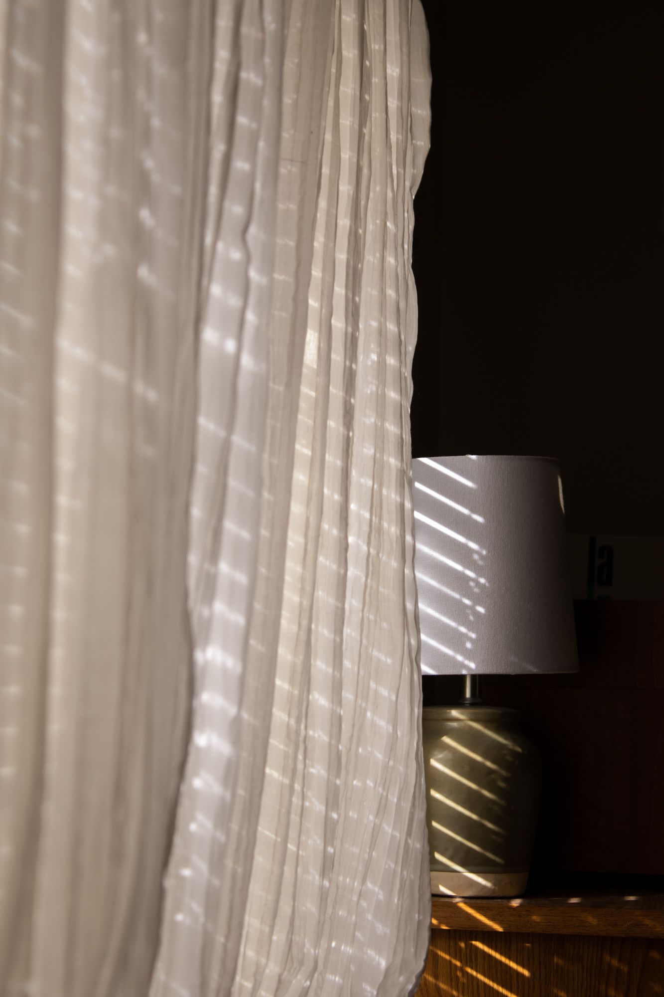 Minimalist interior detail capturing soft sunlight filtered through white curtains, creating a serene, moody play of light and shadow.