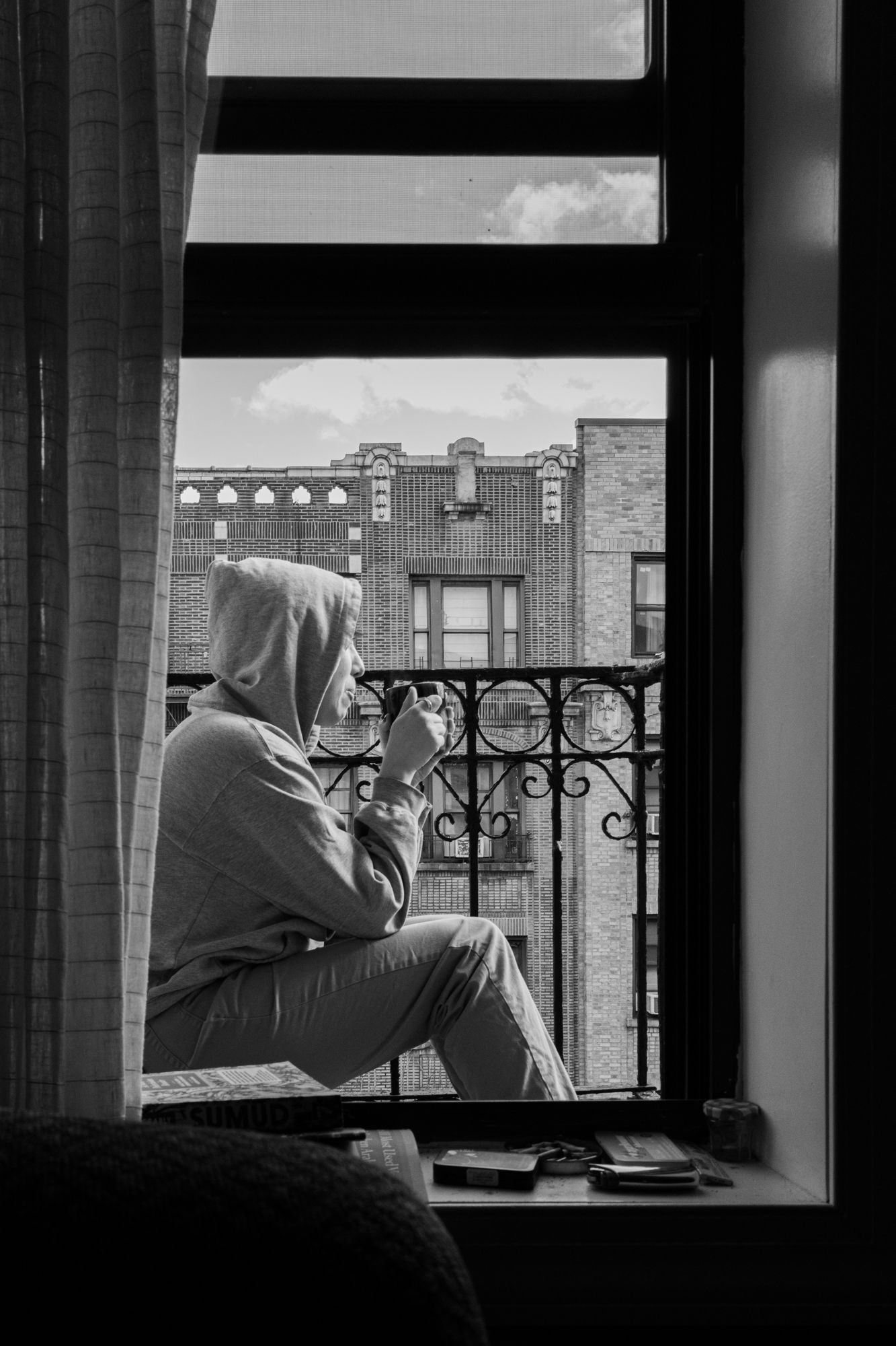 Moody black & white urban lifestyle photography. Silhouette of a person in a hoodie by a window. High-contrast city stock imagery.
