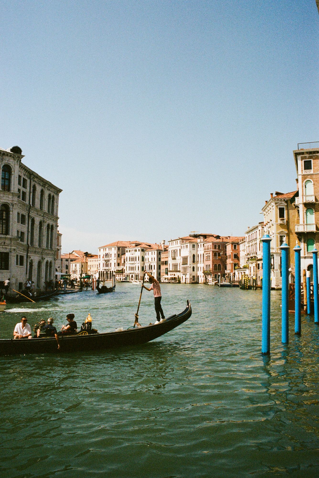 A gondolier navigates the sparkling Grand Canal in Venice, flanked by historic palazzos and iconic blue mooring poles under a clear sky.