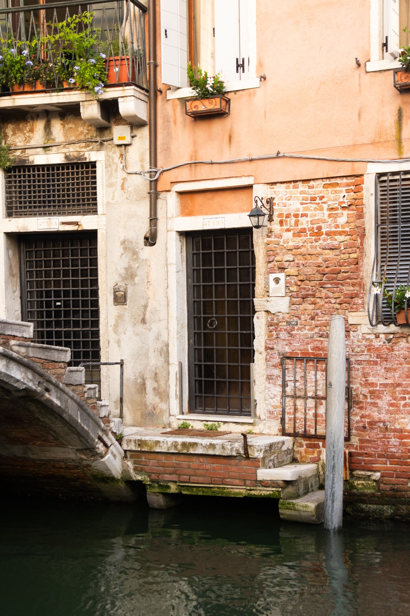 Discover the hidden charm of Venice with this view of a weathered brick facade, iron-grated doors, and stone steps leading to the water.