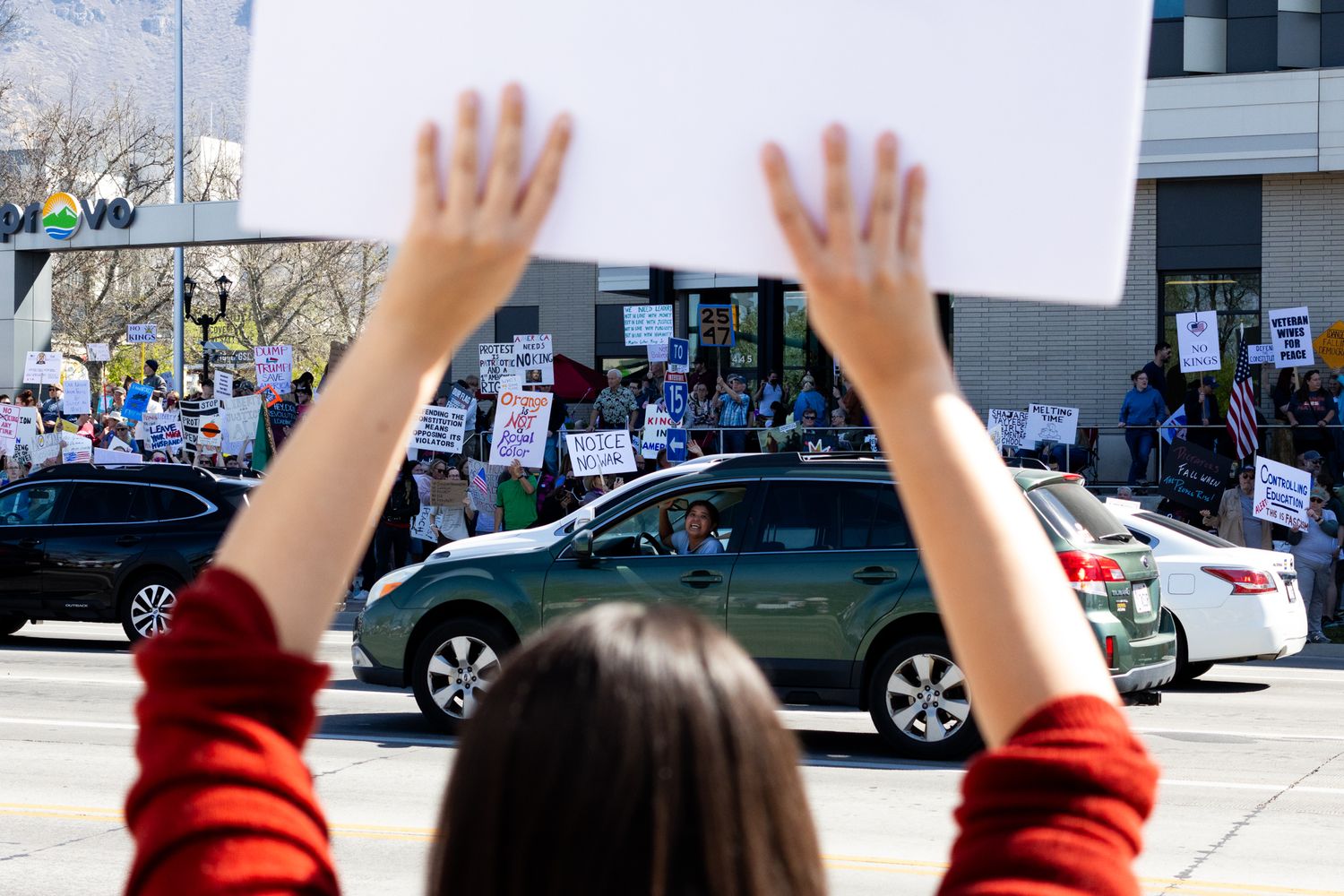 A documentary shot of a protestor holding a blank sign aloft, overlooking a busy street filled with demonstrators and passing cars.