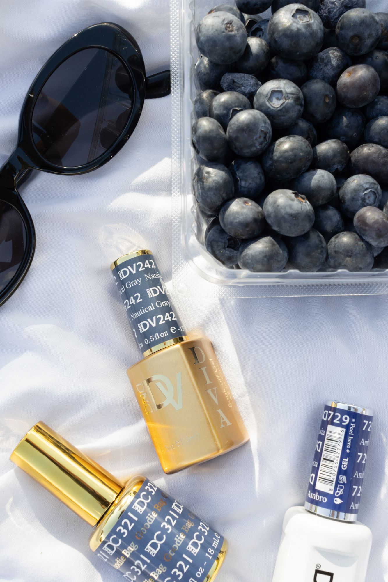Artistic beauty flat-lay with gold-tone nail lacquer, blueberries, and sunglasses, showcasing creative product styling for summer branding.