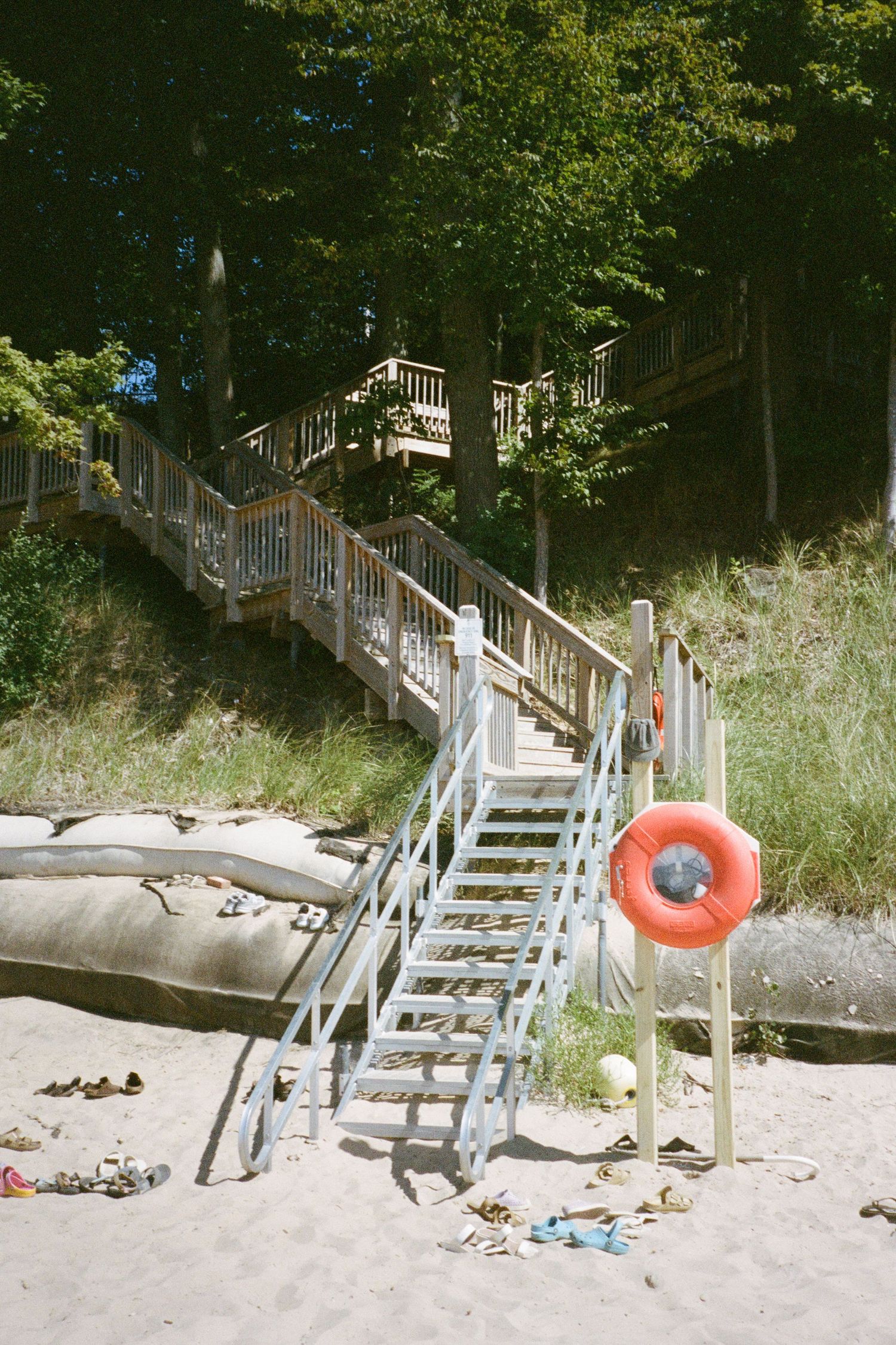A winding wooden staircase leads down to a sandy Lake Michigan beach, complete with a life ring and scattered sandals on a bright summer day