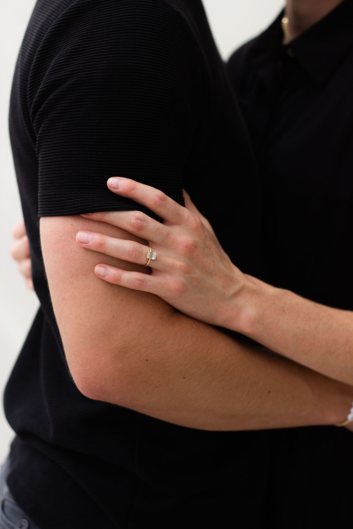 A close-up, intimate shot of a woman’s hand resting on a man’s arm, highlighting an elegant engagement ring against their black outfits.