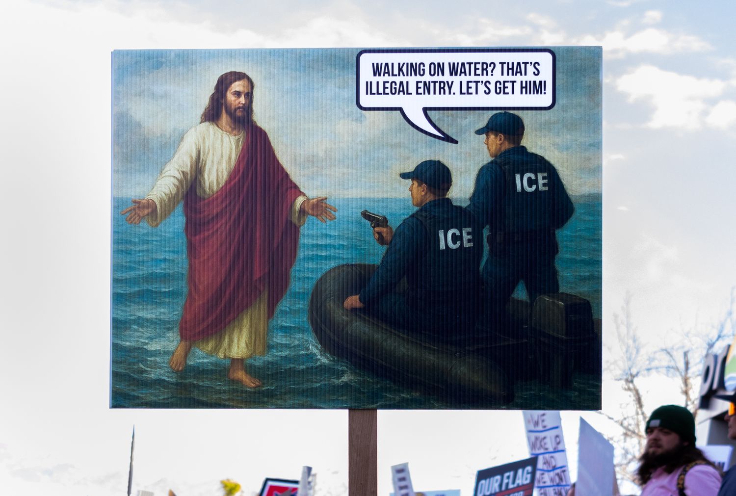 A documentary close-up of a satirical protest sign using religious imagery to comment on immigration enforcement and border policy.