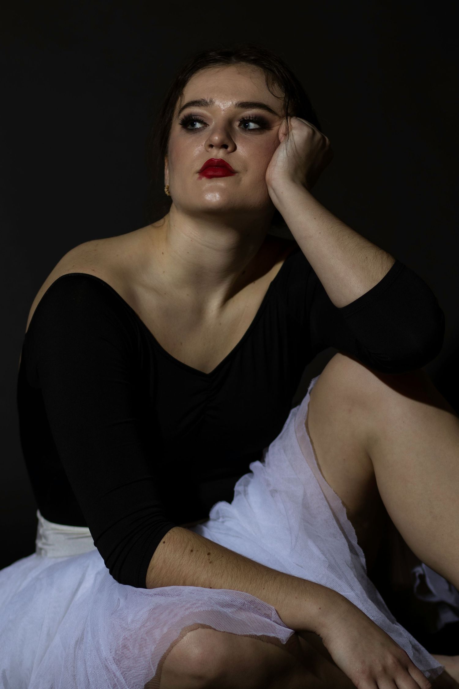 A pensive ballerina in a white tutu and black leotard sits in a dimly lit studio, her thoughtful expression highlighted by dramatic shadows.