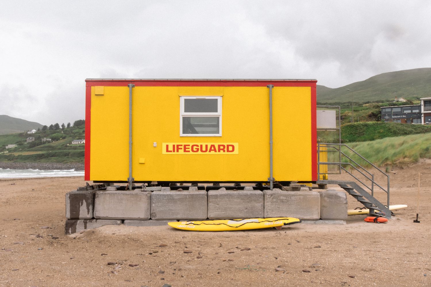 Lifeguard station. Focus on the bright yellow geometry and repetitive concrete blocks against the muted beach. Sharp, industrial rhythm.